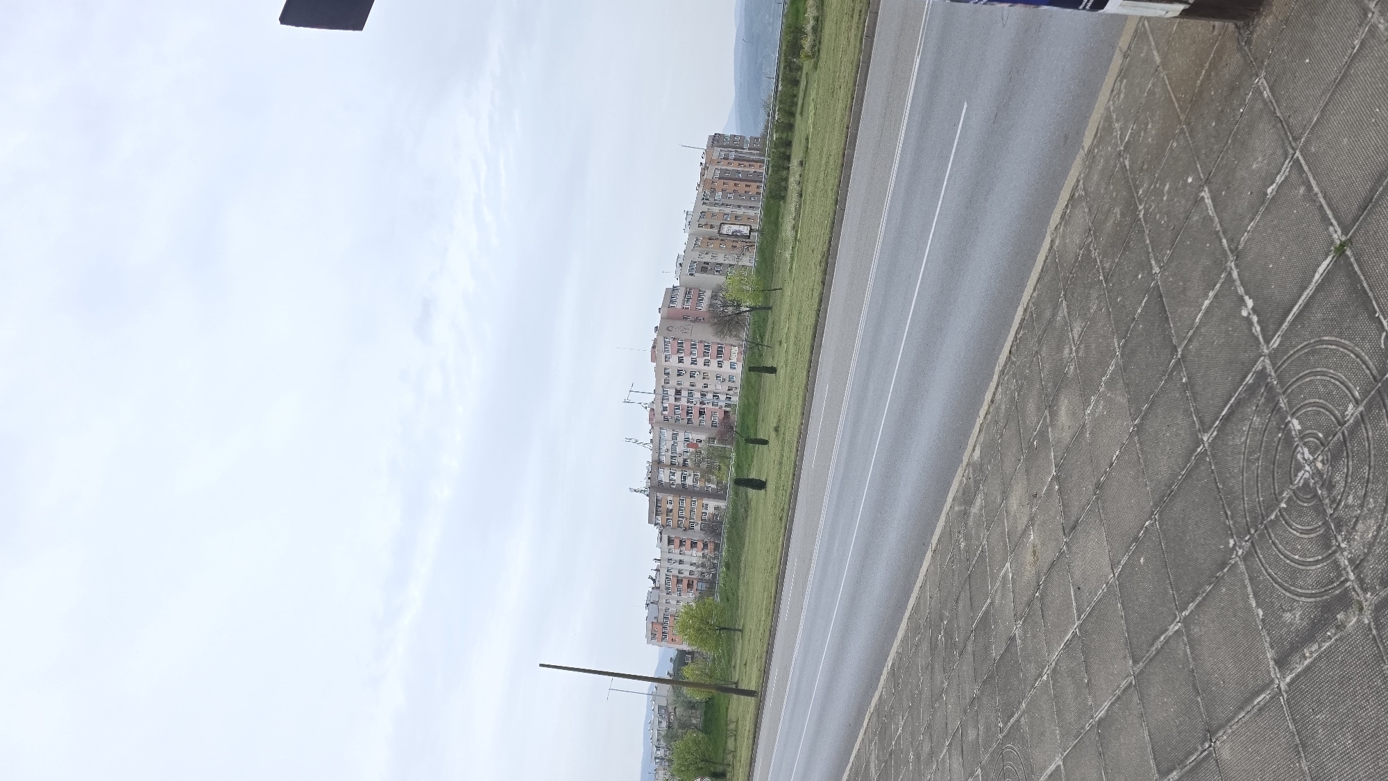The image captures a view of residential buildings in Blagoevgrad, Bulgaria, taken from a street-level perspective. A paved sidewalk with circular patterns is visible on the right side of the frame, adjacent to a multi-lane road with white lane markings. Beyond the road, a strip of green grass separates the road from a row of tall apartment buildings, varying in color from tan to pink. The buildings are densely packed and stretch into the distance, terminating at the edge of the frame. The sky is overcast with light grey clouds, and distant mountains can be seen on the horizon. A dark, rectangular object is visible at the top edge of the frame, potentially a sign or part of a structure. A few thin trees and poles are sporadically placed between the road and buildings. The photograph appears to be taken during the daytime, with soft, diffused light.