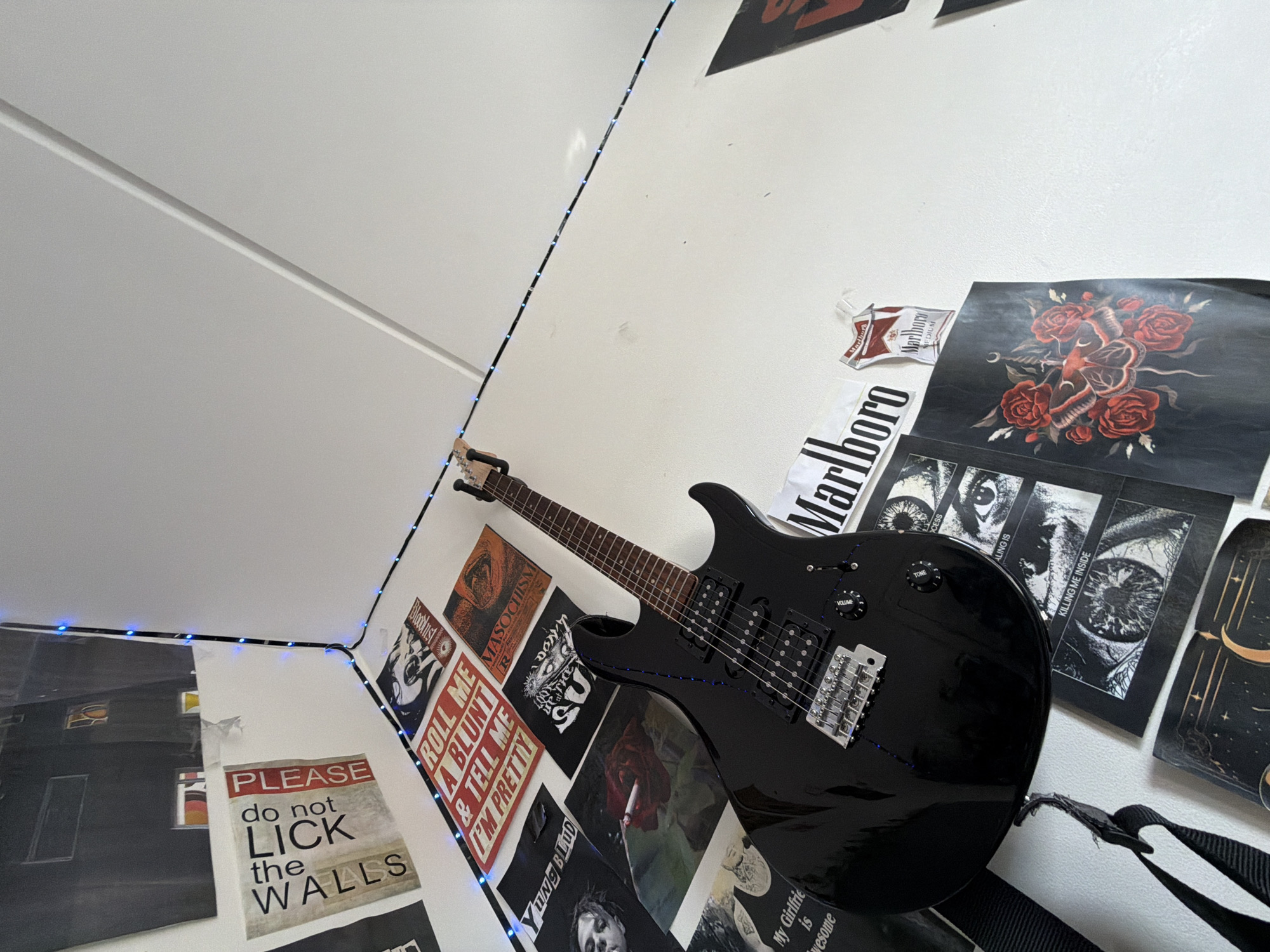 An indoor shot, from a low angle, shows a black electric guitar leaning against a wall. The wall is covered with various posters and decorations, including a "Please do not lick the walls" poster, a "Roll me a blunt & tell me I'm pretty" poster, a "Masochism" poster, posters with eye imagery, a poster of red roses and a moth, and a "Marlboro" cigarette pack. Blue LED string lights are draped along the wall and ceiling. The overall aesthetic appears to be that of a decorated bedroom or personal space. No people are visible in the image. The scene is static.