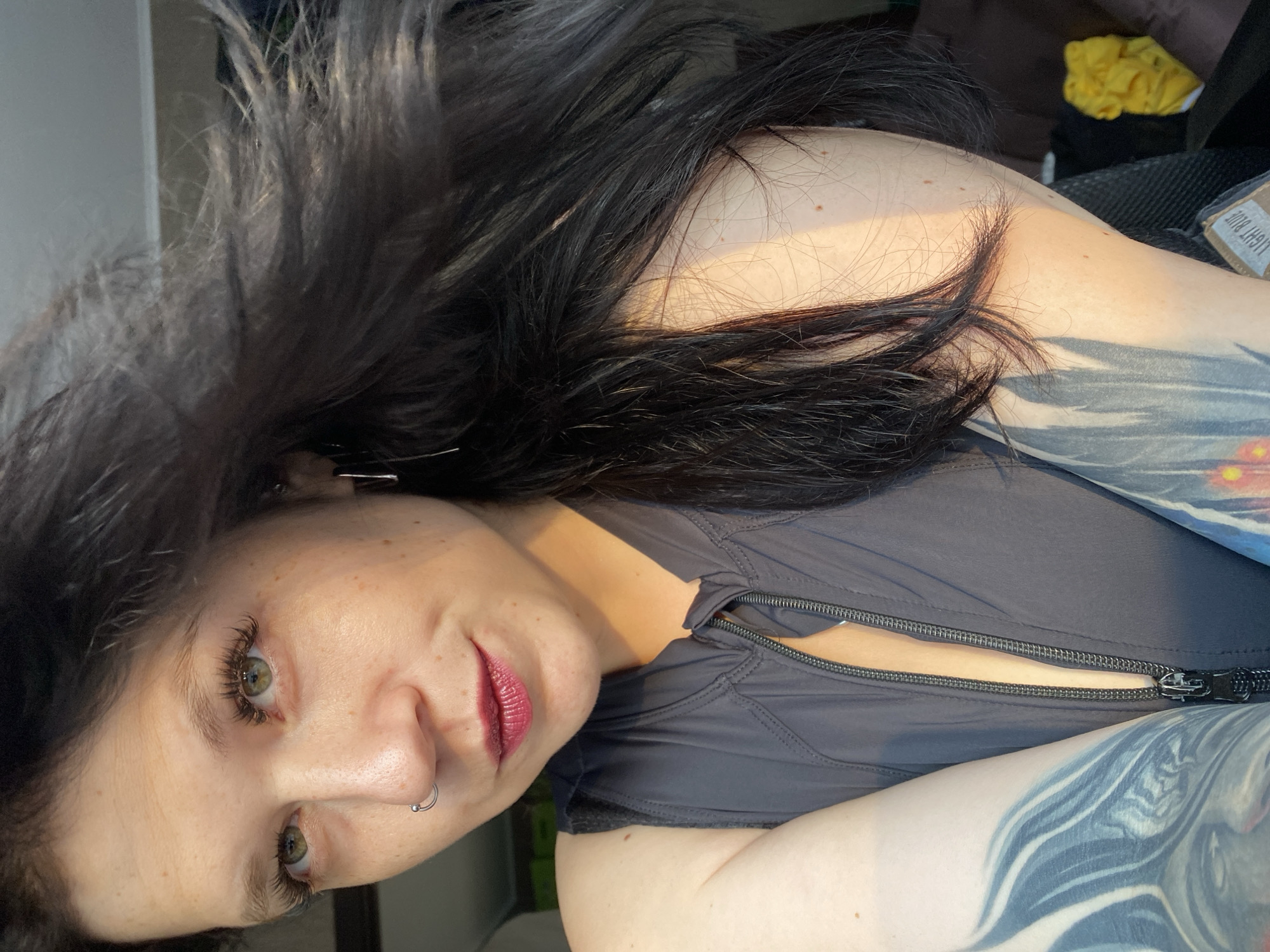 A young woman with long, dark hair and green eyes is pictured in a close-up, portrait-style shot. She has freckles across her nose and cheeks and is wearing dark lipstick. She has a nose ring and an earring visible. Her hair is spread out around her head and shoulders. She is wearing a dark grey, zip-up top. Visible tattoos with intricate designs are on her arm and shoulder. The lighting suggests it is either early morning or late afternoon, with sunlight casting shadows and highlighting parts of her skin. The background is out of focus but appears to be an indoor setting, possibly a room, with some out-of-focus items including a yellow fabric and a tag that reads "LIGHT PURE". The overall mood is relaxed and perhaps contemplative.