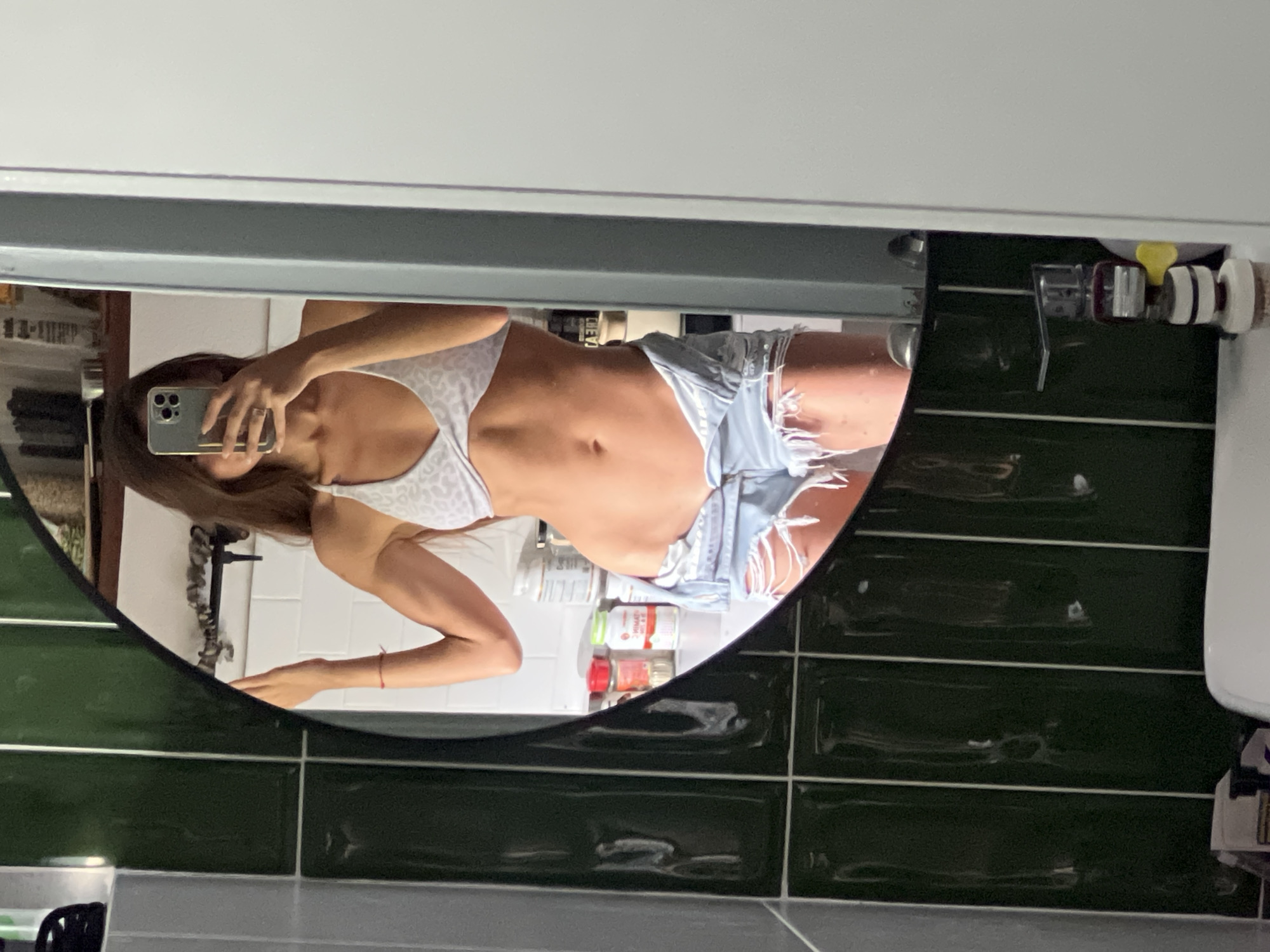 A person, likely female, takes a mirror selfie in an indoor setting, location Unknown, Unknown. The individual is reflected from the chest to mid-thigh in an oval mirror with a black frame. They wear a light-colored, patterned bikini top with a twisted front design and light blue ripped denim shorts, unbuttoned at the waist to expose the abdomen and navel. A smartphone, held in the right hand, obscures the face. A red string bracelet is visible on the left wrist. The background, reflected in the mirror, shows a counter or shelf with several white containers (possibly supplements), smaller bottles with red and green caps (one labeled "VITAMIN"), and a wall tiled with white subway tiles. A wooden cabinet and stacked items (possibly books) are also visible in the reflection. To the left of the mirror, the wall is covered with dark green, glossy rectangular tiles with white grout. To the right, a plain white wall is visible. Below the mirror, a white surface and several small bottles with varied caps are present.