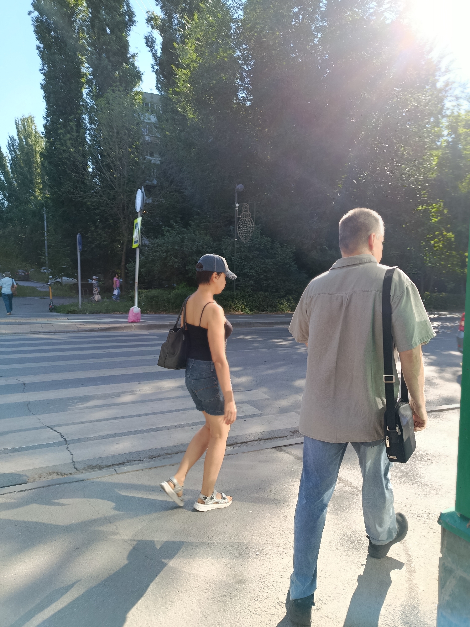 The image shows a sunny day in Saratov, Russia, with two people walking on a crosswalk. In the foreground, a man in a light brown shirt and blue jeans walks away from the camera, carrying a black messenger bag. To his left, a woman in a black tank top and denim shorts, wearing a baseball cap, walks in the same direction. She is also carrying a black handbag. Behind them, several other pedestrians are visible in the distance. The scene is lined with tall trees and a building is visible behind the foliage. The sunlight creates strong shadows on the crosswalk.