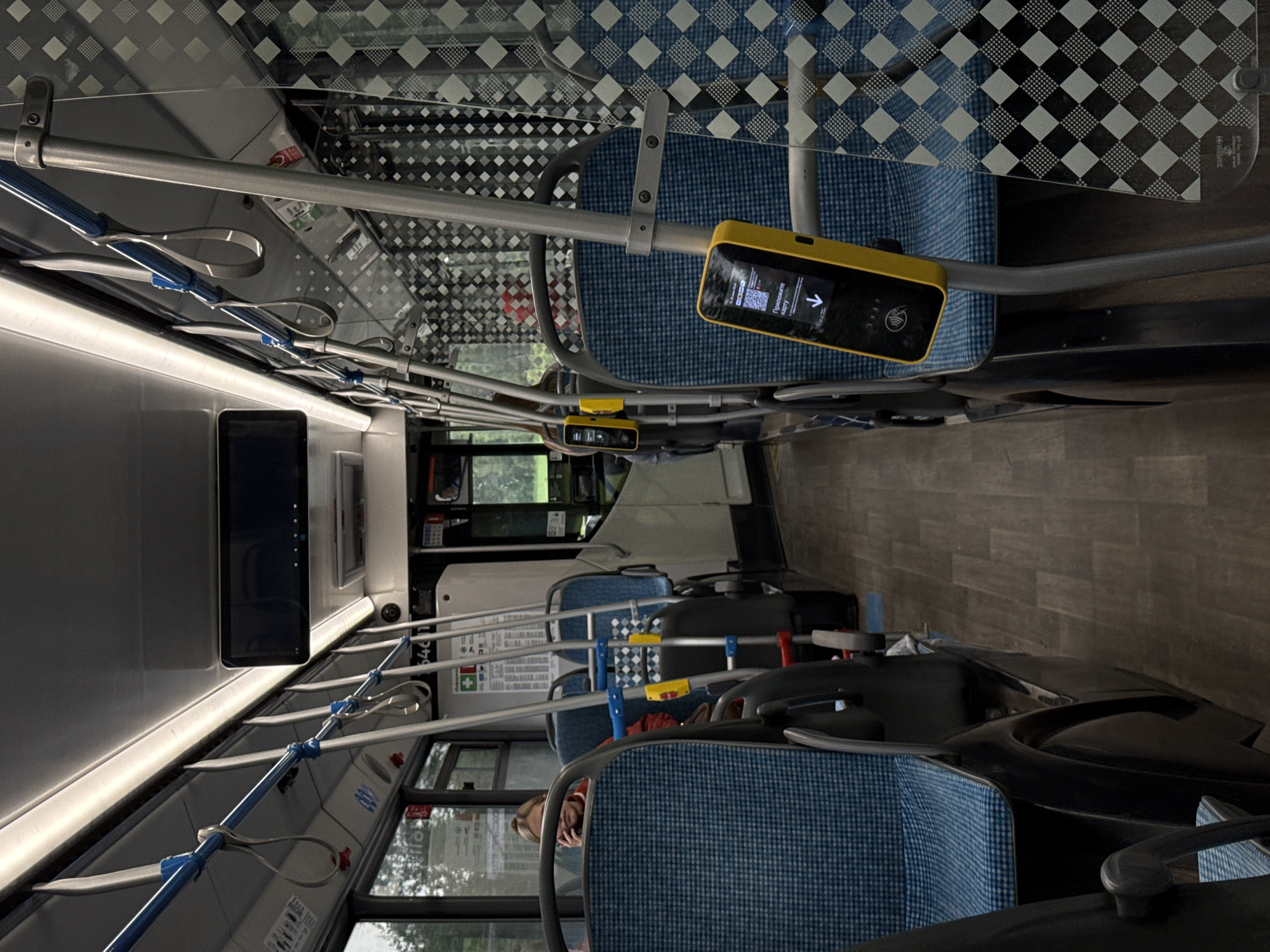 The image shows the interior of a bus in Yakovlevskoye, Russia, looking down the aisle towards the front. The seats are blue with a checkered pattern, and there are metal poles and hand straps for passengers. A woman with blonde hair is visible on the left, talking on her phone. On the right side of the aisle, a yellow card reader is mounted on a pole. The floor is dark wood-grain.
