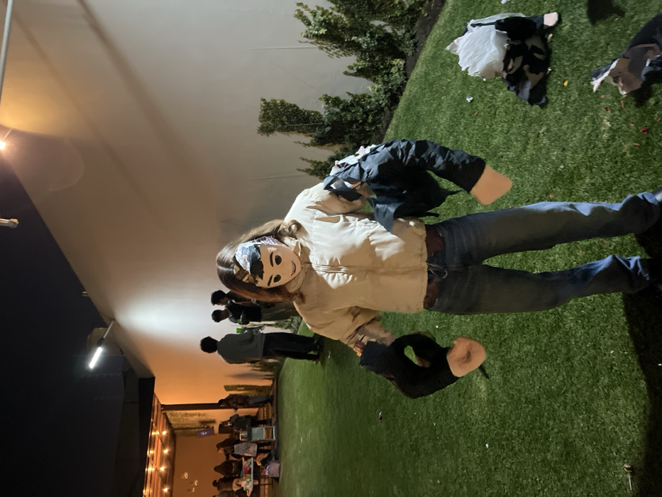 A person stands on green grass, wearing jeans, a tan jacket, and a mask. Next to them is a torn-up piñata. In the background, other people are seated at tables, and some are standing nearby. A white wall runs along one side, illuminated by lights. A bush or hedge is behind the person. The scene seems to be an outdoor gathering or party at dusk.