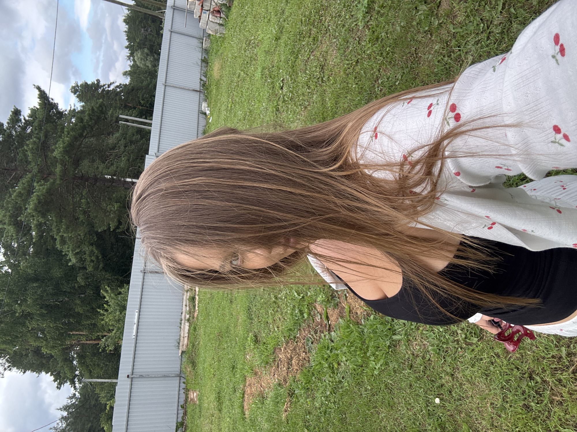 A young woman with long, light brown hair is standing in a grassy yard, possibly a backyard or a rural area in Прикамский, Russia. Her hair is partially obscuring her face as she looks towards the camera. She is wearing a black tank top and a white cardigan with a cherry pattern. In her left hand, she holds a red object, possibly a hair accessory or a small tool. Behind her, there is a grey corrugated metal fence and trees in the background. The ground is covered in green grass with some patches of dry earth. The sky is cloudy. The overall impression is a casual, outdoor setting.