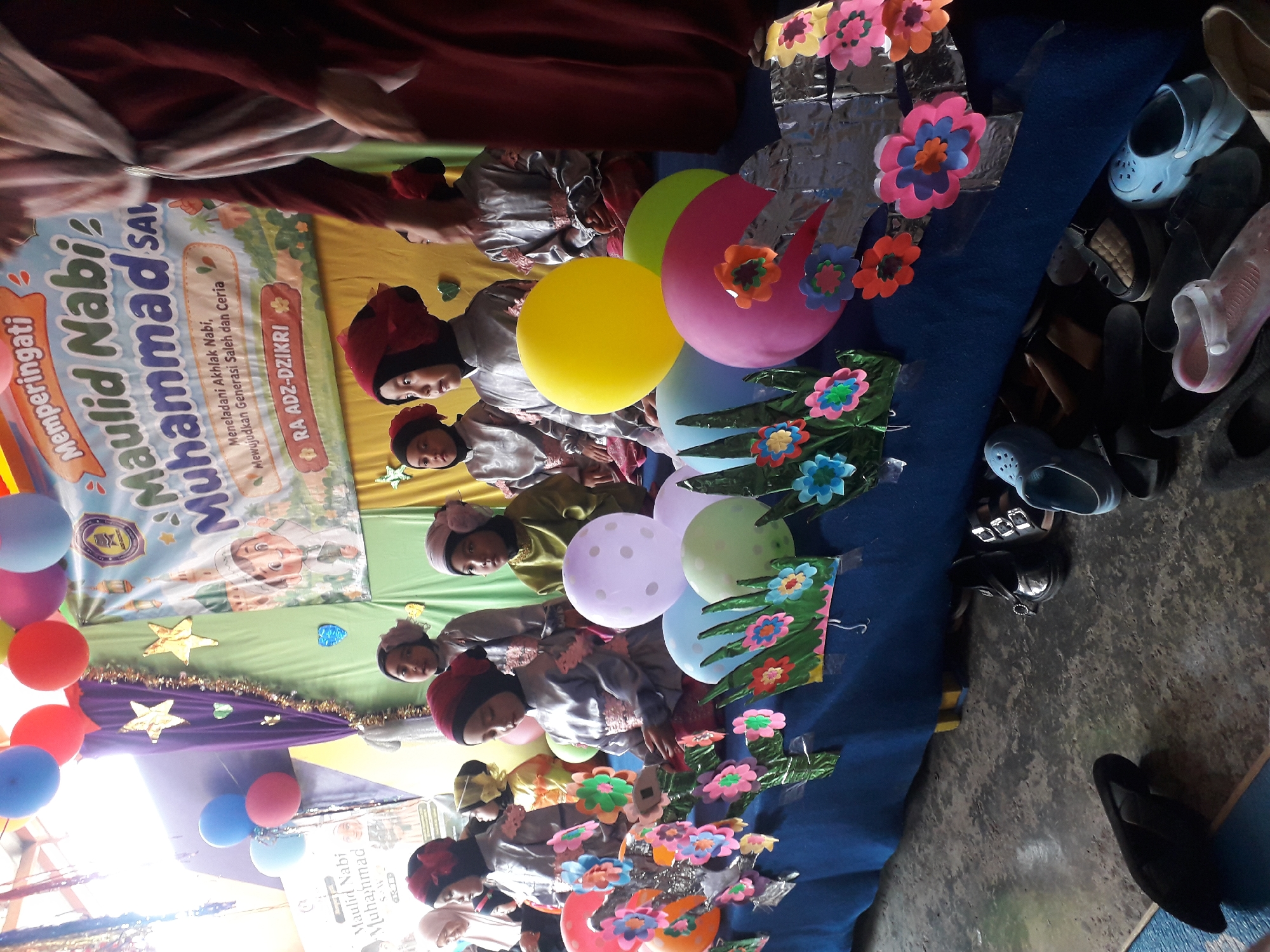 This image shows a group of children, likely students, dressed in formal attire and participating in an event. They are standing behind a table decorated with colorful balloons, paper flowers, and tinsel. A banner in the background reads "Memperingati Maulid Nabi Muhammad SAW" (Commemorating the Birthday of the Prophet Muhammad SAW), indicating a religious celebration. The children are wearing traditional Islamic clothing, with some wearing hijabs and others wearing kufis. The overall atmosphere appears festive and celebratory. The event is likely taking place in Cankuang, Indonesia, given the context of a Maulid Nabi celebration. The children are attentive, possibly performing or listening to a speaker. The presence of a teacher or adult in the background, dressed in maroon, suggests an organized event within a school or community setting. The ground is concrete, suggesting an indoor or semi-outdoor venue. Various shoes are visible on the floor, indicating that people may have removed their footwear before entering the area.