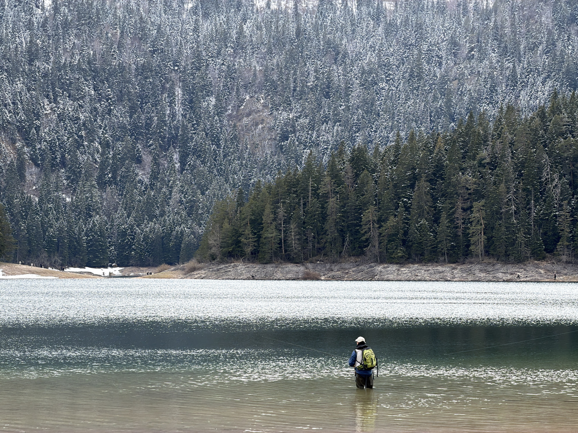 The image presents a serene winter or early spring landscape of a lake in Pitomine, Montenegro. In the foreground, a lone person stands waist-deep in the calm, shallow water, facing away from the viewer and engaged in fishing. They are dressed in dark waders, a blue jacket, a light-colored cap, and a distinctive lime-green backpack, and are holding a fishing rod with a line extending into the water.
The lake's surface, rippled by a gentle breeze, shimmers with light, indicating daytime. The water near the shore is a light, somewhat sandy brown, gradually transitioning to a deeper green-blue in the distance.
The background is dominated by a dense evergreen forest covering a steep mountain slope. The upper parts of the forest are heavily dusted with snow, highlighting the cold conditions, while the trees closer to the lake on the opposite bank appear darker green with less snow. The distant shoreline features patches of exposed earth and residual snow. Several small, indistinct figures can be observed along the far bank, suggesting other people are present, possibly walking or observing the scene. The overall atmosphere is one of tranquility and natural beauty. No text is visible in the image.