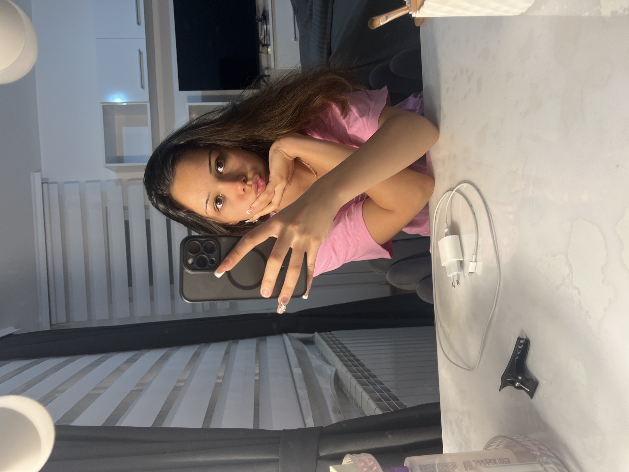 A young woman with long, dark brown hair, wearing a pink t-shirt, is taking a selfie in what appears to be an indoor setting. She looks directly into the camera with a playful pout, her right hand resting near her chin, while her left hand holds a dark-colored smartphone. Her nails are manicured with white French tips, and her ring finger features a small decorative embellishment.
She is situated at a light-colored (white/off-white) table or vanity. On the table to her right, a white phone charger (adapter and cable) and a black hair clip are visible. The background shows a room with various elements: a dark sofa or bed with cushions in the upper right, and white wall-mounted shelves on the upper left. A partial view of a round white light fixture is seen on the far left. A large reflective surface, likely a mirror, shows a reflection of vertical window blinds and other parts of the room, including the back of her phone. The overall lighting suggests an indoor setting, possibly with artificial light, rather than bright daylight. The image itself is oriented sideways, rotated 90 degrees clockwise from a standard portrait view. There is no discernible text in the image.