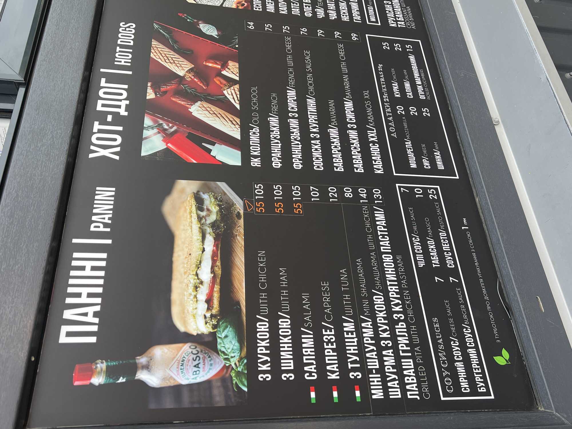 This is a close-up photograph of a food menu board displayed outdoors. The menu is divided into two main sections: "ПАНІНІ / PANINI" and "ХОТ-ДОГ / HOT DOGS."
The left side of the board showcases panini options with corresponding prices in what appears to be Ukrainian currency. The panini choices include variations with chicken, ham, salami, caprese, and tuna. There are also mini-shawarma and grilled pita options listed. Below the panini section, a selection of sauces is offered, including chili sauce, Tabasco, cheese sauce, and pesto sauce, each with a listed price.
The right side of the board features hot dog options. These include varieties such as "Як колись / Old School," "Французький / French," and "Французький з сиром / French with cheese," as well as sausages and "Баварський / Bavarian" variations.
Add-ons ("Додатки 25r / Extras 25g") are listed at the bottom, including mozzarella, chicken, salami, cheese, ham, and pickled cucumbers.
The board has a black background with white text. Images of the food items are displayed alongside the menu descriptions. The presence of Ukrainian text indicates that the menu is likely located in Ukraine. Based on other evidence of Ukrainian writing elsewhere in the prompt images, it is reasonable to believe that this menu is located in Ternopil, Ukraine.