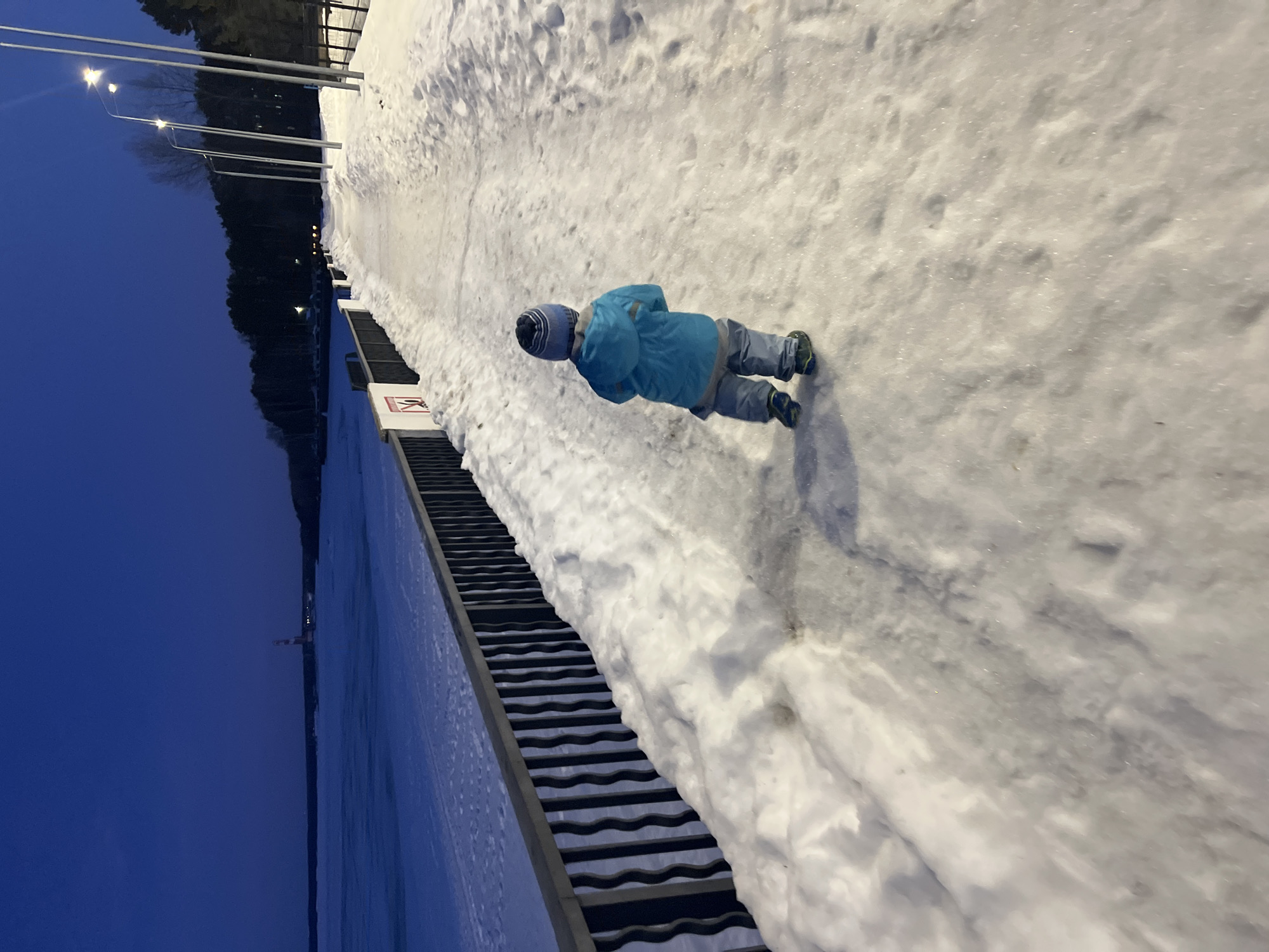The image shows an outdoor winter scene at dusk or night, on the embankment in Chaikovsky, Russia, likely along the Votkinsk Reservoir. A single young child, viewed from behind, is walking on a snow and ice-covered paved pathway that extends into the distance, curving slightly right. The child wears a blue and white striped beanie, a bright blue hooded jacket, light grey pants, and dark shoes with green accents.
To the child's left, a dark metal railing with vertical, wavy balusters separates the pathway from a vast, frozen body of water covered in snow, reflecting the deep blue sky. A white sign with a red 'no entry' or 'caution' symbol is affixed to a post on the railing. In the far distance across the frozen water, a red and white striped lighthouse is visible, along with a dark treeline.
To the child's right, a row of tall, thin streetlights illuminates the pathway and surrounding snow with yellowish-white light. Behind the streetlights and extending into the background is a dense treeline or forest. Snowbanks line both sides of the pathway, which shows signs of foot traffic. The sky is a deep, consistent blue.
