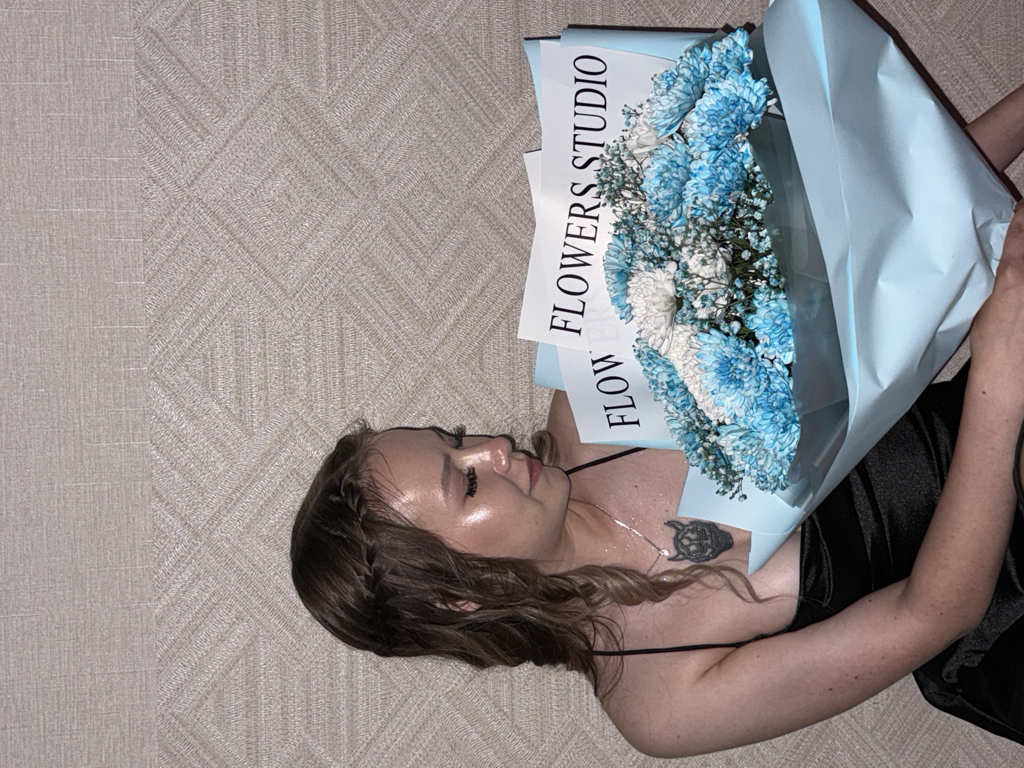 A young woman with blonde hair styled in braids is holding a bouquet of blue and white flowers. She is wearing a black dress and a necklace with a pendant. The background is a textured wall. The image does not contain any information that would allow for determination of location in the city of Chaikovsky, Russia.