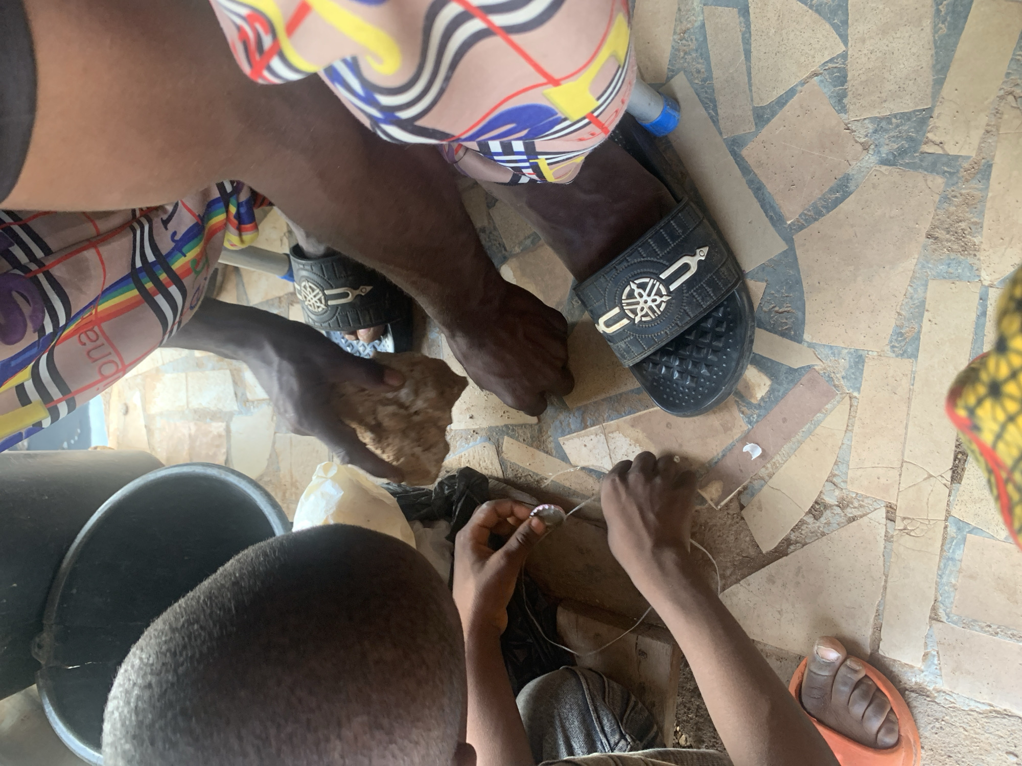 This top-down photograph captures a group of people engaged in a collaborative activity in Sabon Tasha, Nigeria, within what appears to be an indoor setting with an irregularly tiled floor. The floor consists of various shapes of beige and light brown tiles interspersed with grey grout.
At least three individuals are present. In the upper portion of the frame, a person with dark skin is seen wearing a colorful, patterned shirt featuring yellow, red, blue, and black stripes on a beige background, with the word "buona" partially visible. This individual also wears black slides with intricate metallic emblems. Their legs and lower torso are visible.
To the left, the crown of a person's head with short, dark hair is visible, alongside two dark, cylindrical buckets. This person's hands are holding a light-colored, roughly textured object, possibly a food item or raw material. A white plastic bag and another white, amorphous item are also near the buckets.
In the lower center and right, another person's hands are meticulously working on a small, round, metallic object, which appears to be a bottle cap or similar item, using a thin white string or thread. This suggests a craft like beading, jewelry making, or an assembly task. A dark piece of fabric or plastic lies beneath their hands. This individual wears light-colored pants and an orange sandal is visible on their foot.
The scene suggests focused, manual work taking place during daylight. No strong emotions are discernible, but the posture and actions indicate concentration. A small blue-capped object, possibly a pen or marker, is visible near one of the black slides.