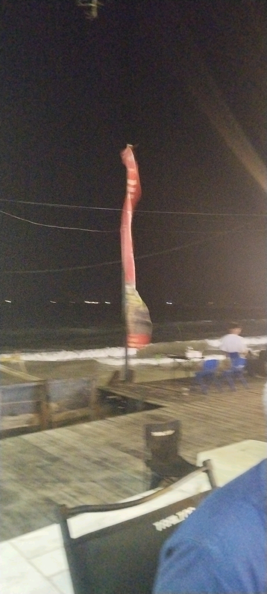 An outdoor scene at night depicts a wooden deck area with a beach and ocean in the background. A tall, thin, red and white striped flag or banner is prominently displayed, appearing to be a sail-like structure, and is attached to a pole. The deck is furnished with several blue chairs and a trash bin. In the background, blurred figures are visible, including one person seated and another standing. The overall impression is that of a beachside establishment. The image is grainy and out of focus, indicating a low-quality capture or a moving camera. The location is identified as Balikpapan, Indonesia.