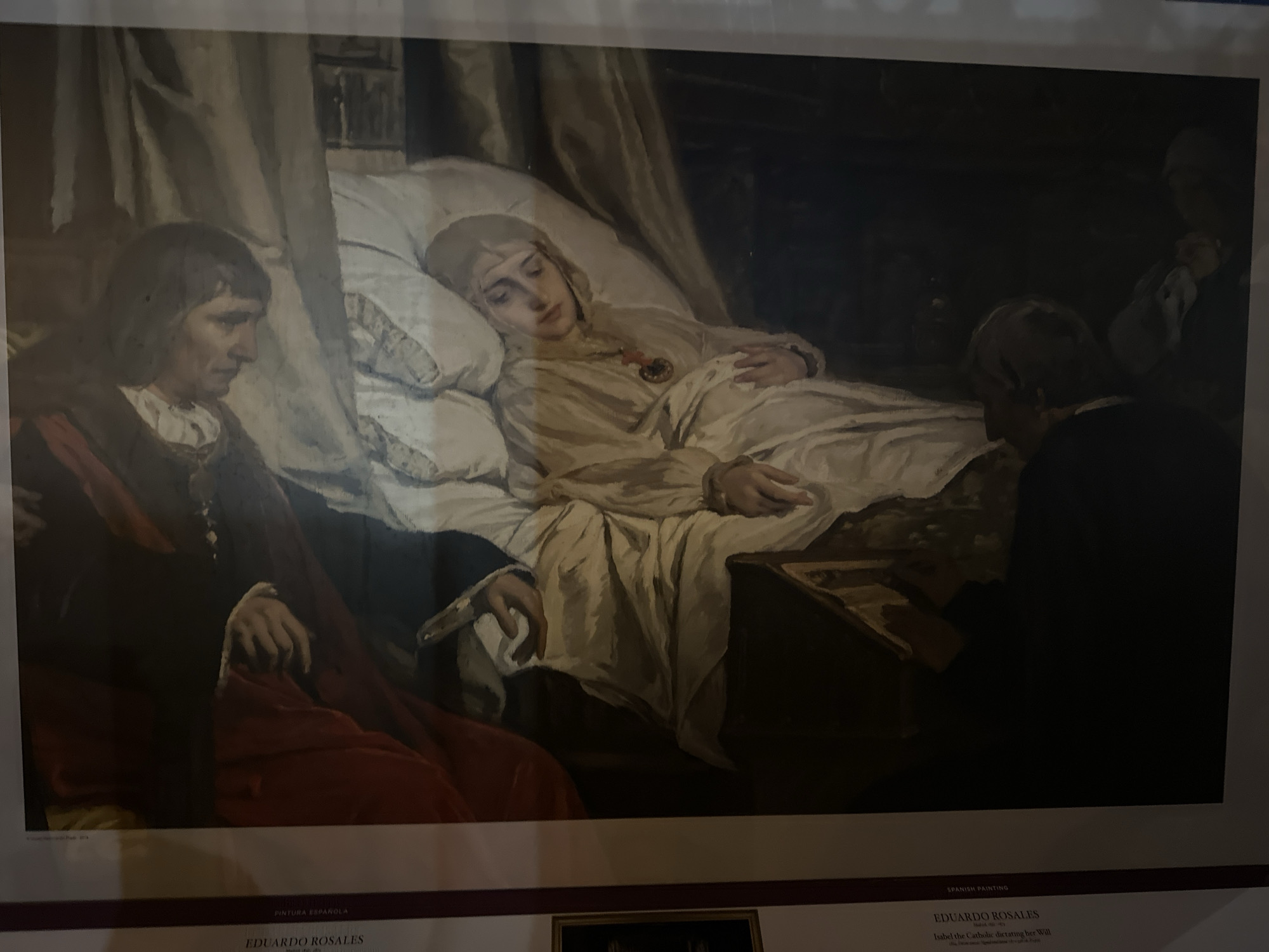 The image shows a painting of a woman lying in bed, appearing ill. She is wearing a light-colored gown and a head covering, with a medal around her neck. Two figures are attending to her. On the left, a person in dark clothing with red fabric draped over their shoulder looks down at the woman. On the right, another person, partially obscured by darkness, leans over a table next to the bed. The setting appears to be a dimly lit room with a canopy over the bed. Below the painting, text identifies the artwork as "Isabel the Catholic dictating her Will" by Eduardo Rosales. The text also indicates "SPANISH PAINTING" and mentions the city of Mazarrón, Spain, suggesting the painting may be displayed there.