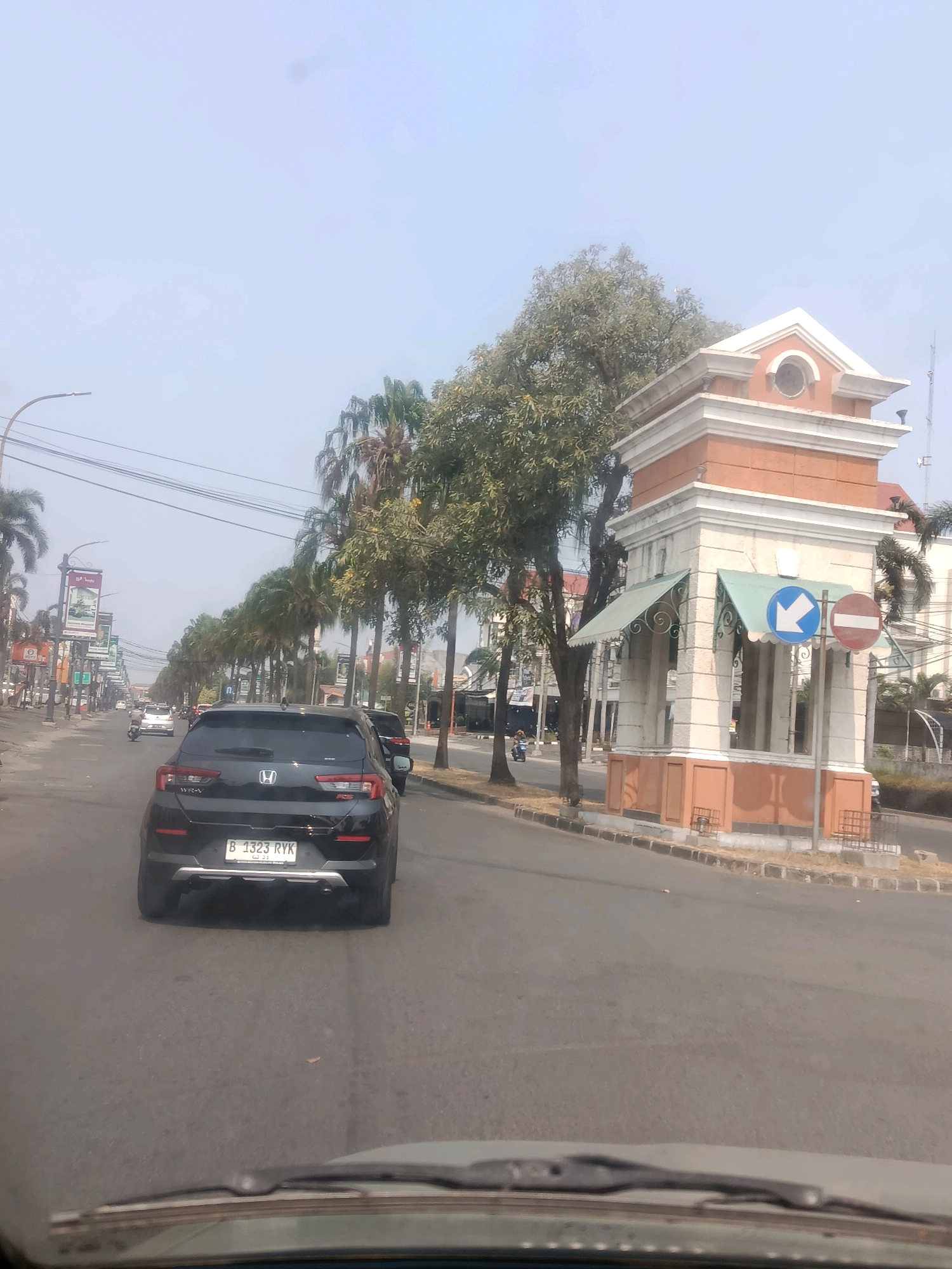 The image depicts a street scene in Bekasi, Indonesia, on a clear, sunny day. In the foreground, the windshield and dashboard of a vehicle are visible, suggesting the image was taken from inside a car. The road ahead is lined with palm trees and other foliage. On the right side of the road is a decorative tower-like structure with an orange and white facade, featuring arched openings and awnings. Two traffic signs are mounted on a pole near this structure: a blue sign with a white arrow pointing left, and a circular red and white sign indicating "no entry" or a prohibition.
Further down the street, several cars are in motion, including a black SUV in the immediate foreground with a license plate reading "B 1323 RYK". A motorcycle is also visible in the distance. Billboards and shopfronts line the left side of the road. The overall atmosphere appears to be that of a typical urban environment in Indonesia.