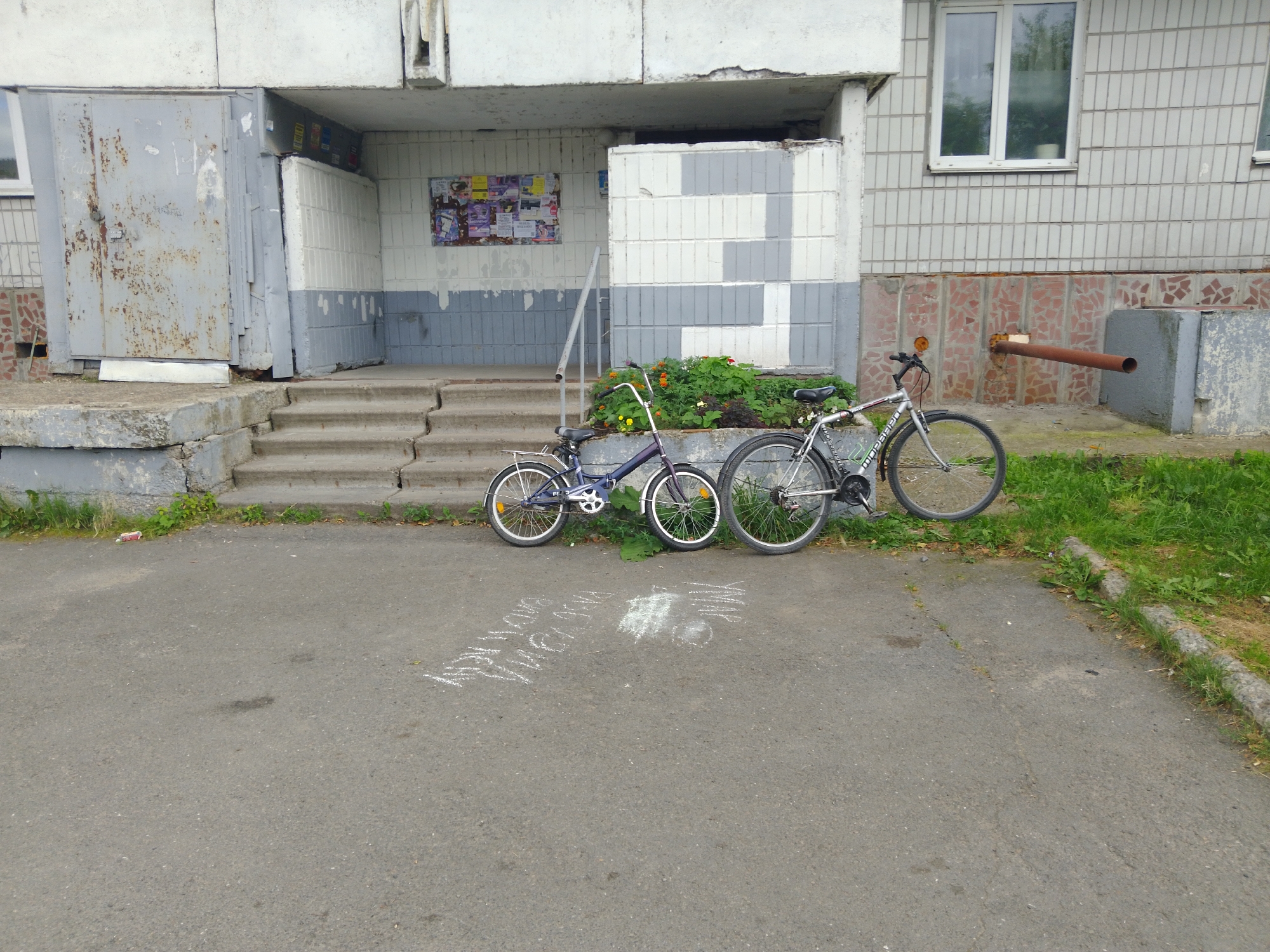 Two bicycles are parked on a paved area in front of a building. The bicycle on the left is smaller and purple, while the bicycle on the right is larger and silver. Both bicycles have their kickstands down. There are chalk drawings on the pavement in front of the bicycles. The building appears to be a multi-story apartment building with a gray facade and a few windows. There is a set of concrete stairs leading up to an entrance with a large metal door. A small flower bed with green plants and some red and yellow flowers is located near the entrance. The area is surrounded by grass.