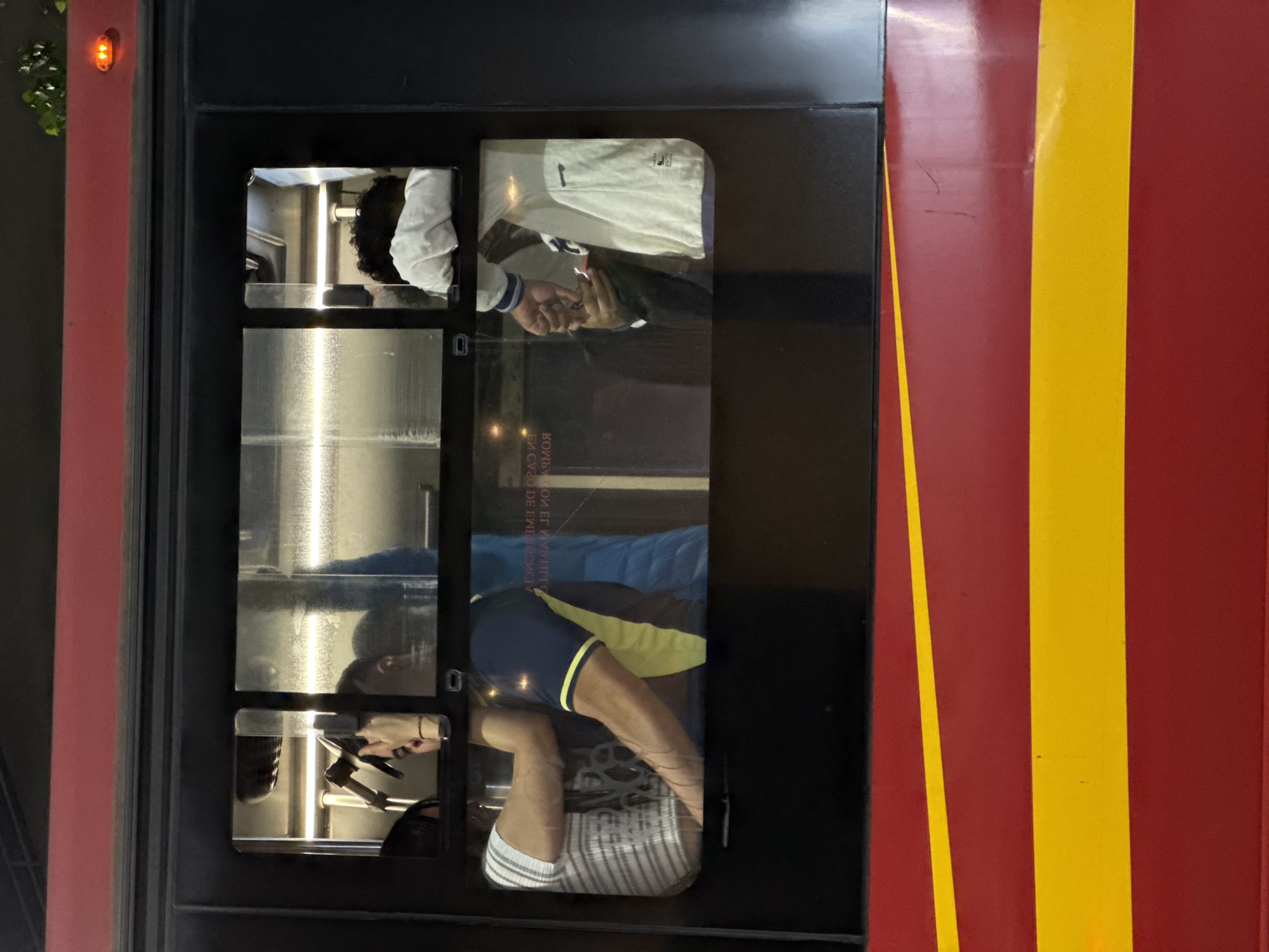 The image depicts a window display, possibly of a laundromat, on a red and yellow striped wall. The display consists of four small windows showing different stages of laundry being done, with people interacting with the laundry in each. The windows are stacked vertically. The location is likely in Mexico City, Mexico.