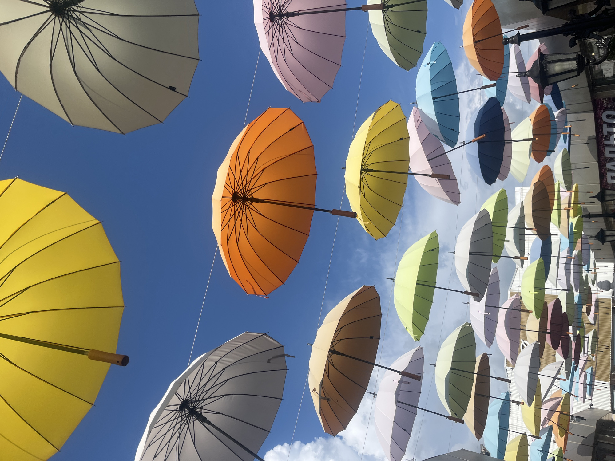 The image depicts a street in Sochi, Russia, adorned with a colorful canopy of open umbrellas suspended in the sky against a clear blue backdrop with scattered white clouds. The umbrellas are arranged in multiple layers, creating a vibrant and whimsical display. They vary in color, including yellow, orange, pink, purple, green, light blue, dark blue, and off-white. The umbrella handles are visible, many made of wood. Below the umbrellas, the upper portions of buildings and some streetlights are visible. The overall scene suggests a festive or decorative installation designed to create a visually appealing urban environment. No people are visible in the image.