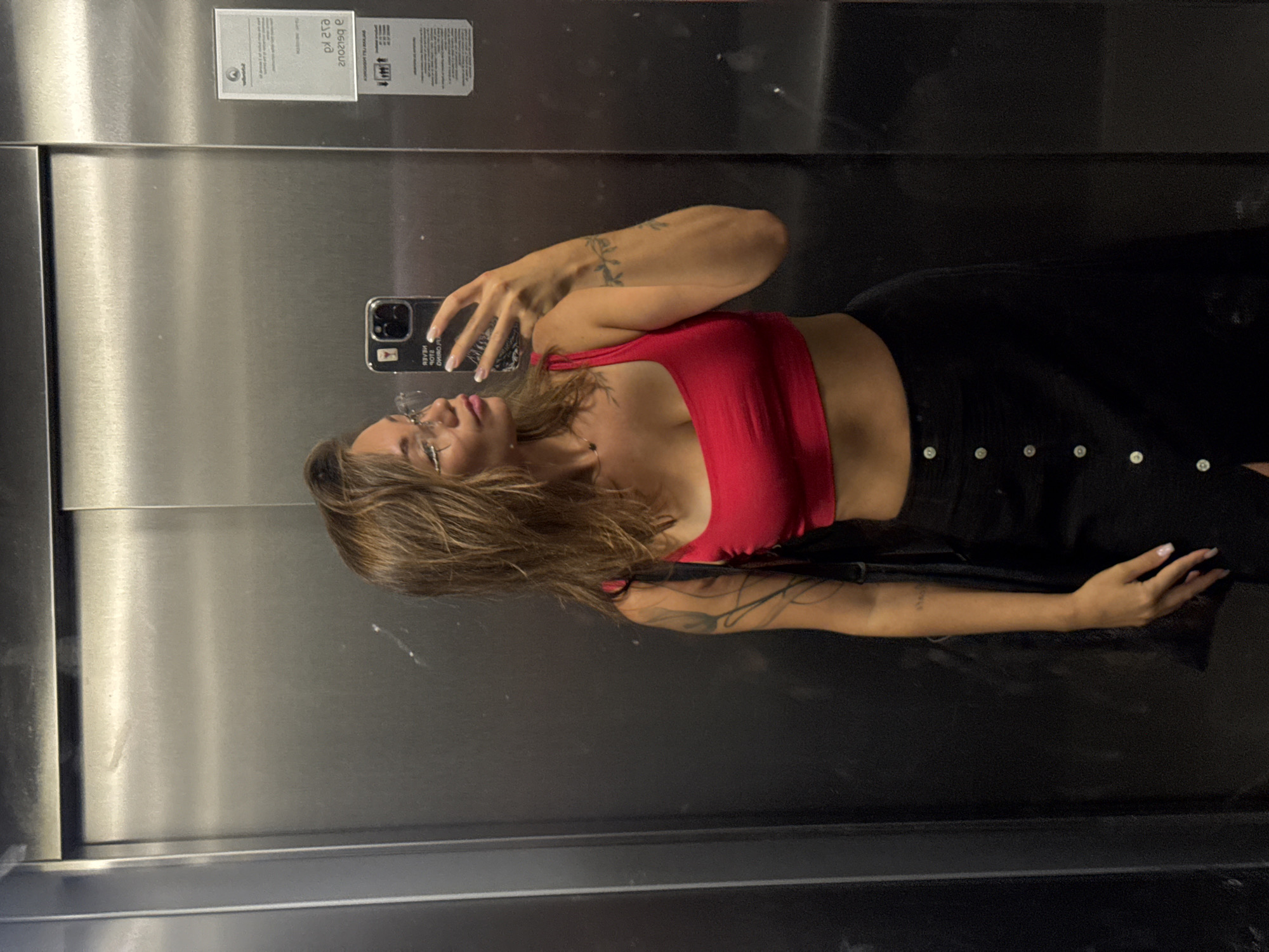 The image depicts a young woman taking a mirror selfie in what appears to be an elevator. She is wearing a red crop top and a black skirt with buttons. She has glasses on and her hair is down. The elevator walls are made of stainless steel. A sticker with text is visible on the right wall of the elevator. Based on the original image and provided hints, the location is likely within the city of Budva, Montenegro.
