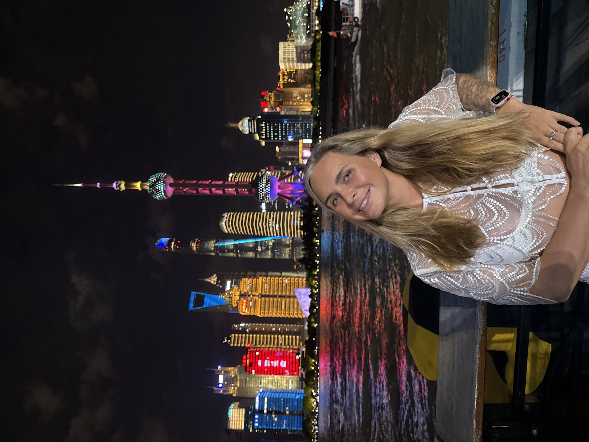 A medium shot captures a woman with blonde hair and a white, sheer, lace top posing for a photo at night. She is leaning on a railing with a body of water in front of her, reflecting the vibrant lights of a cityscape in the background. The skyline is dominated by numerous skyscrapers, with the Oriental Pearl Tower, a prominent landmark, illuminated with pink and purple lights. A boat is visible in the water to the right. The woman is smiling at the camera, and a smartwatch is visible on her left wrist. The scene is set against a dark sky with scattered clouds.