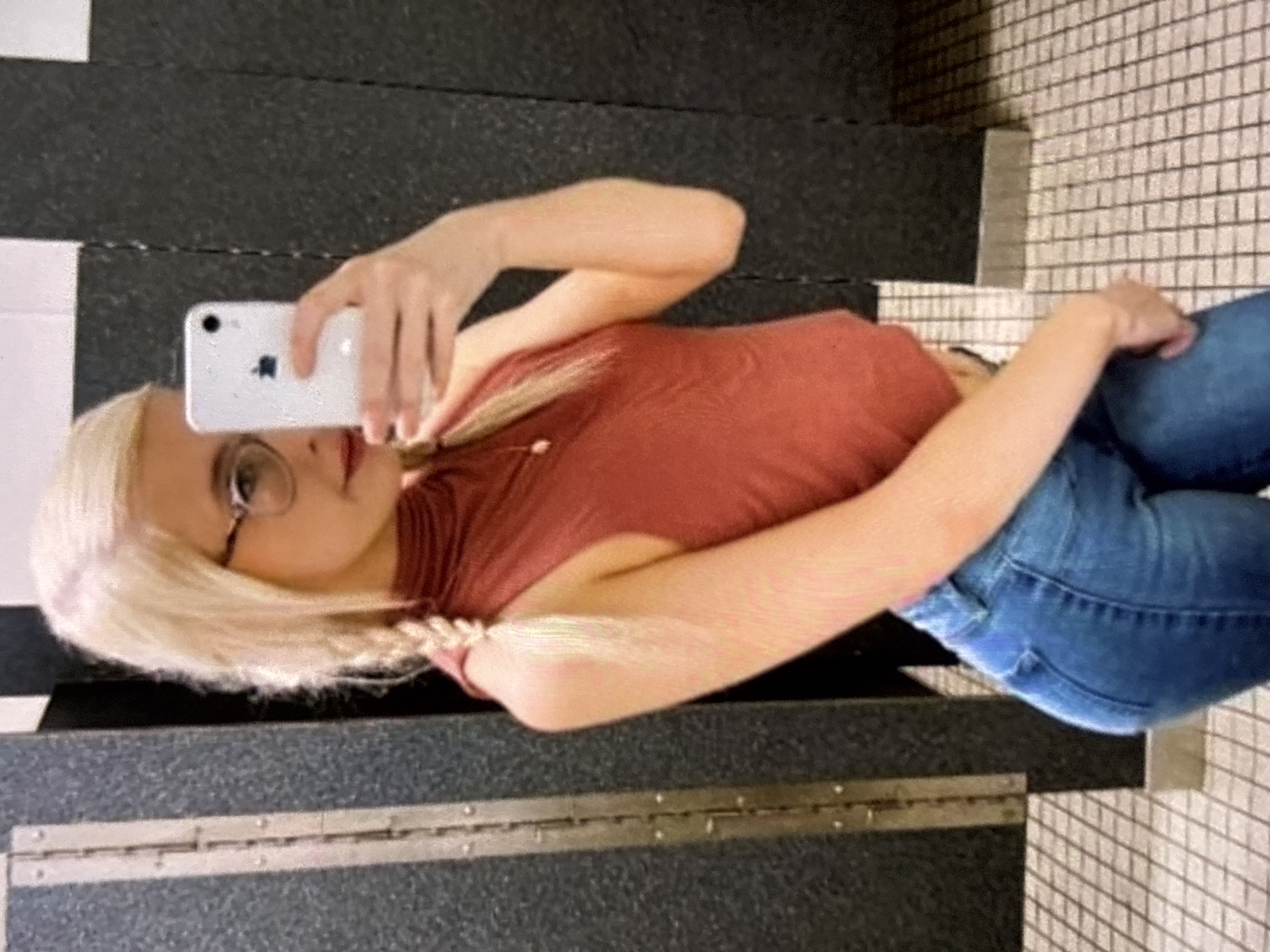 A young woman with blonde hair styled in braids, wearing glasses, a rust-colored sleeveless top, and blue jeans, stands in a bathroom and takes a selfie with a smartphone. She is facing a mirror, with her reflection visible. The bathroom appears to have tiled walls and floors, with stall doors visible in the background.