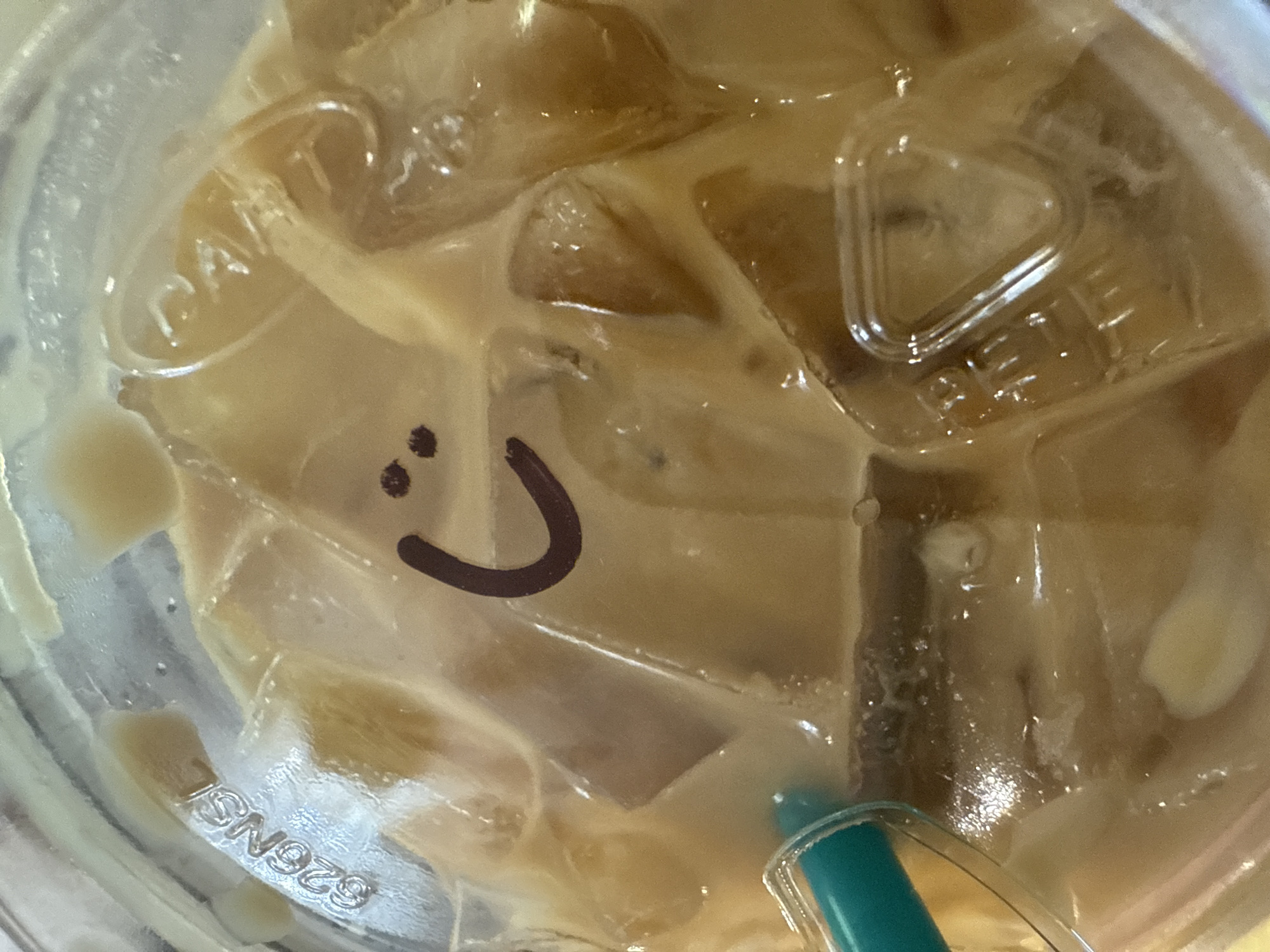A close-up overhead shot depicts a disposable plastic cup filled with a light brown liquid and ice cubes, likely a coffee-based beverage. A green and clear plastic straw is partially visible in the lower right. A crude, drawn-on smiley face in black is present on the plastic lid. Text, "626NSL," is faintly visible embossed on the lower left. The image was taken indoors. There is no information to suggest the location in Davie, United States of America.