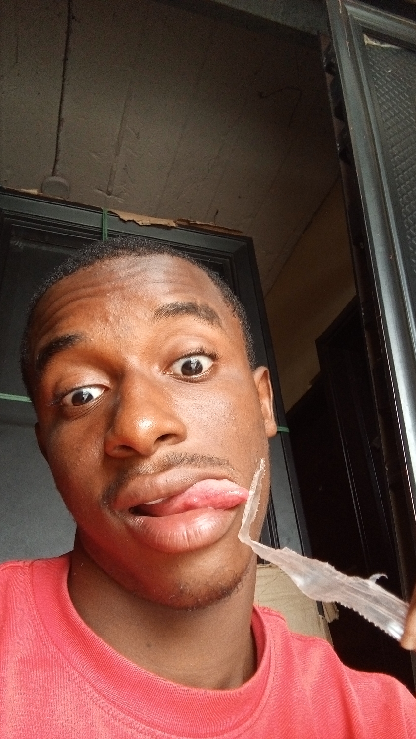 A close-up, eye-level shot captures a young Black man indoors, likely in Nkpor, Nigeria, making a playful expression. He is positioned centrally, looking directly at the camera with wide, surprised eyes. His tongue is extended out of his mouth, touching or positioned very close to a transparent plastic fork that he holds up to the side of his mouth.
The man has short, dark hair, a light mustache, and a small goatee. He is wearing a red or coral-colored crew-neck t-shirt. The lighting suggests it is daytime.
The background is dark and features elements of an interior space. To the left, a dark door frame is visible, with a textured, possibly painted, wall above it that shows some minor wear or peeling. A second dark door frame is partially visible on the far right. A thin green cord or string can be seen stretching horizontally in the upper left portion of the background. There is no visible text in the image.