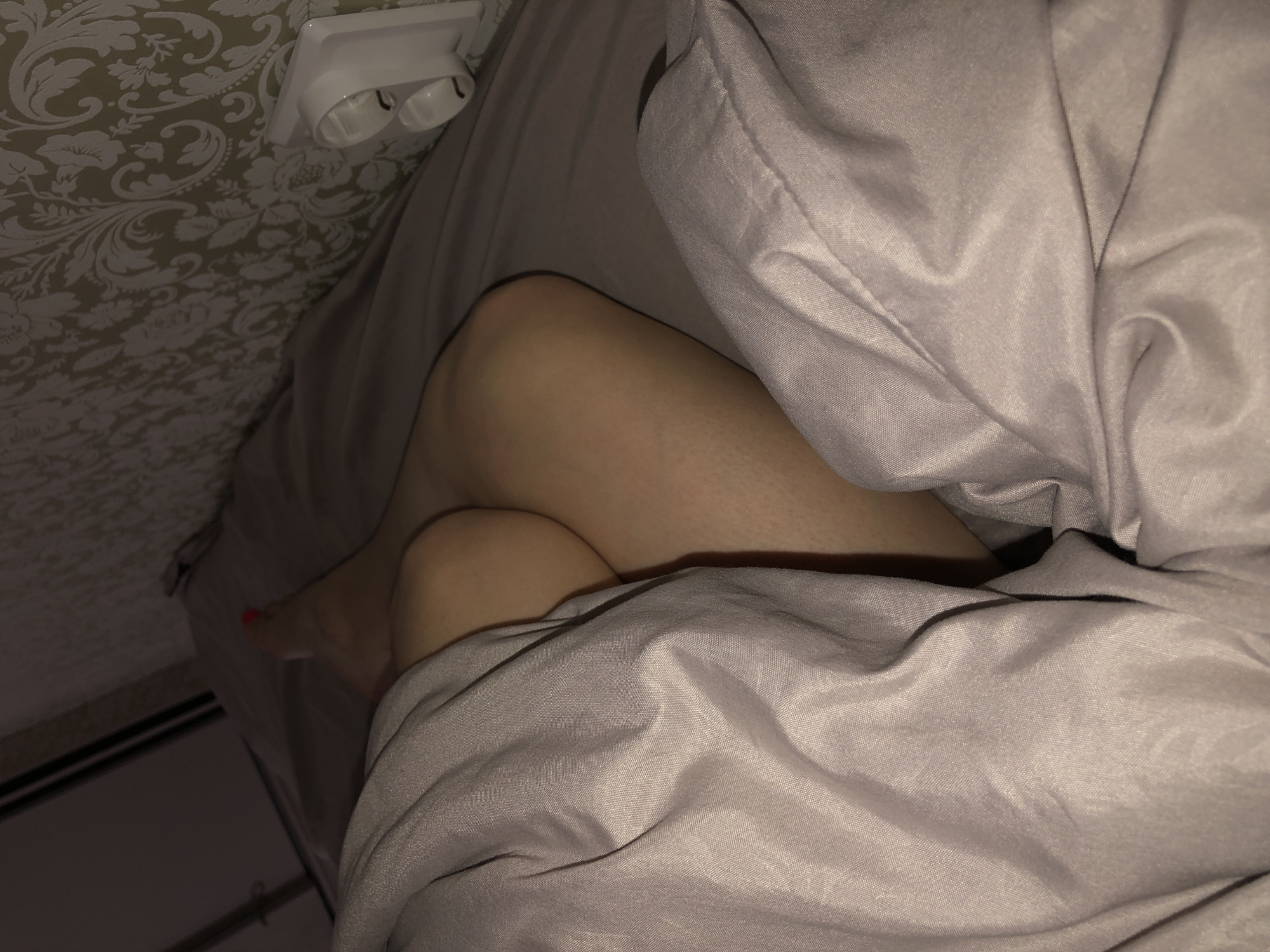 The image shows a person in a bed covered with gray bedding. The person's legs are visible, bent at the knees. One foot with red nail polish is also visible. The background includes a wall with patterned wallpaper and a white electrical outlet. The location in the city of Noviy, Russia is unknown.