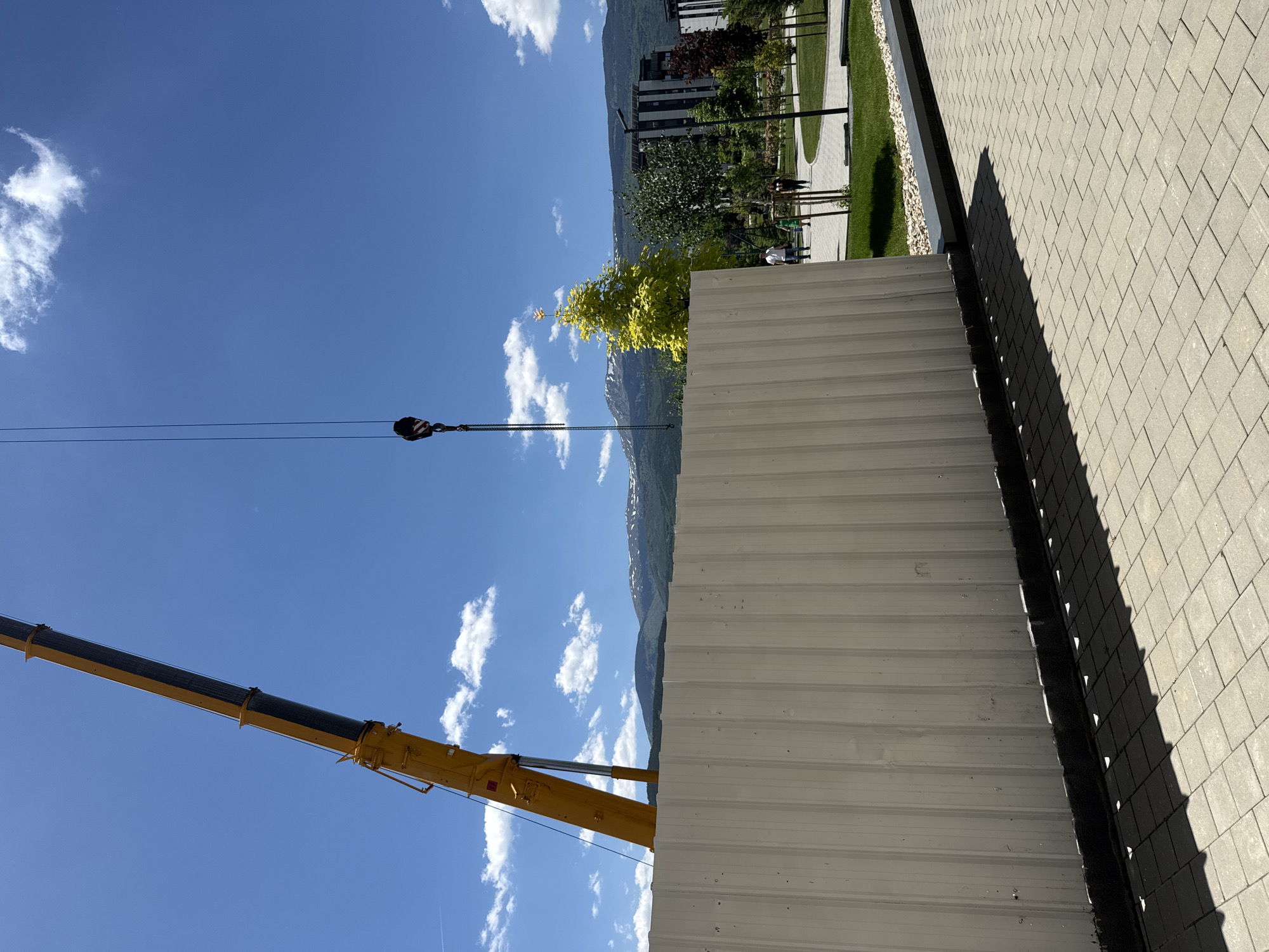 The image depicts a construction scene. A yellow crane's boom extends into the sky. A cable hangs from the crane's hook. Adjacent to the construction area is a paved walkway near a grassy area with trees and a modern building in the background. Mountains are visible in the distance. The sky is blue with scattered clouds. A large metal wall or structure is the immediate foreground element. The location appears to be in an urban or suburban area. This cannot be verified as occurring in Sofia, Bulgaria.