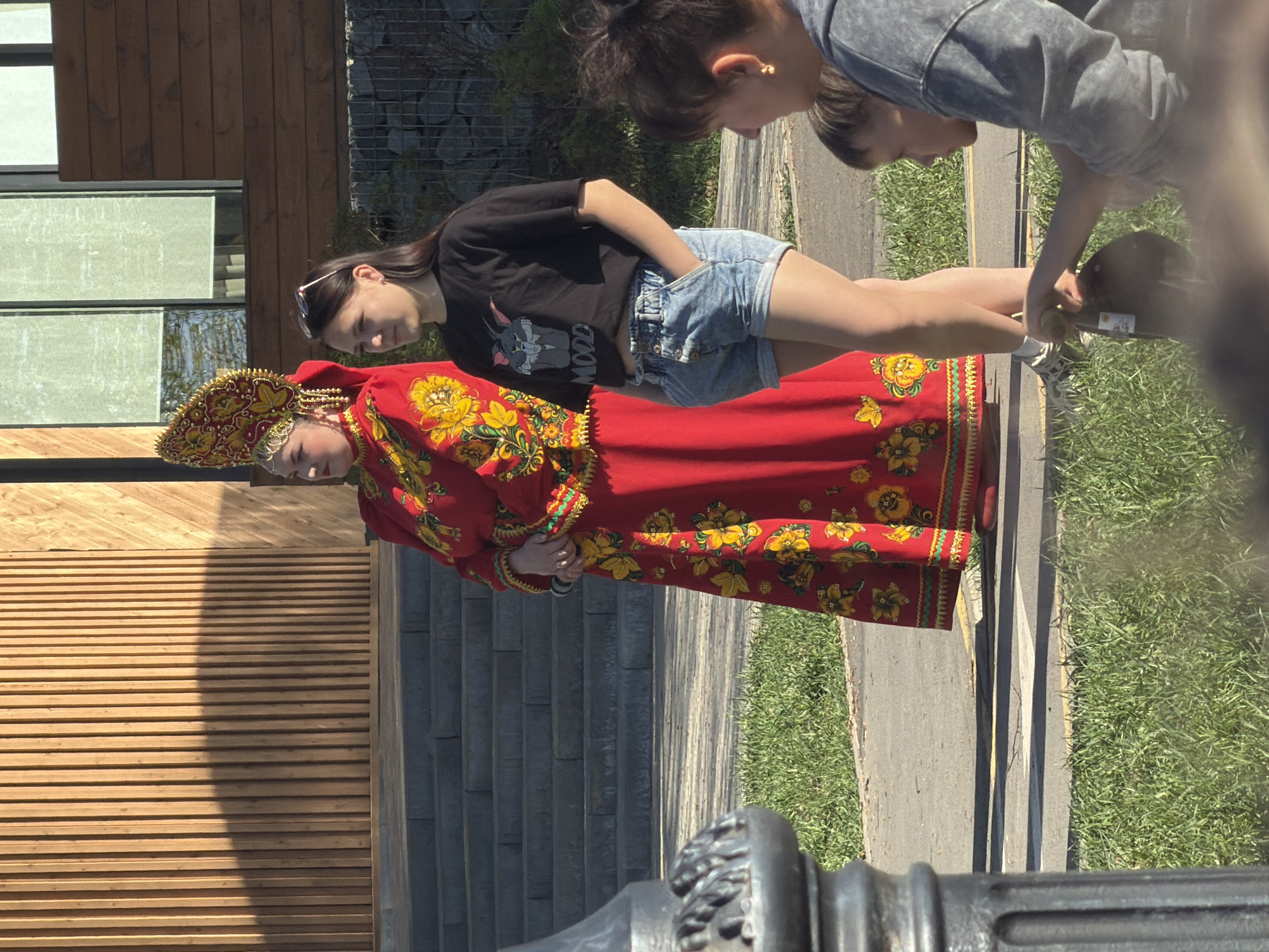 The image shows three people in an outdoor setting. One woman is wearing a traditional Russian dress with a red base and yellow floral patterns, along with a matching headdress. A second person, an adolescent, is wearing denim shorts and a black t-shirt with an animal print. The third person, partially visible, is wearing a gray sweatshirt. The three are located next to a building made of stone and wooden planks, and the image was taken on an asphalt sidewalk next to some grass. A blurry foreground object makes the picture seem to be taken through a barrier. It's difficult to determine the precise location within Votkinsk without further information, but the architecture suggests a public space.