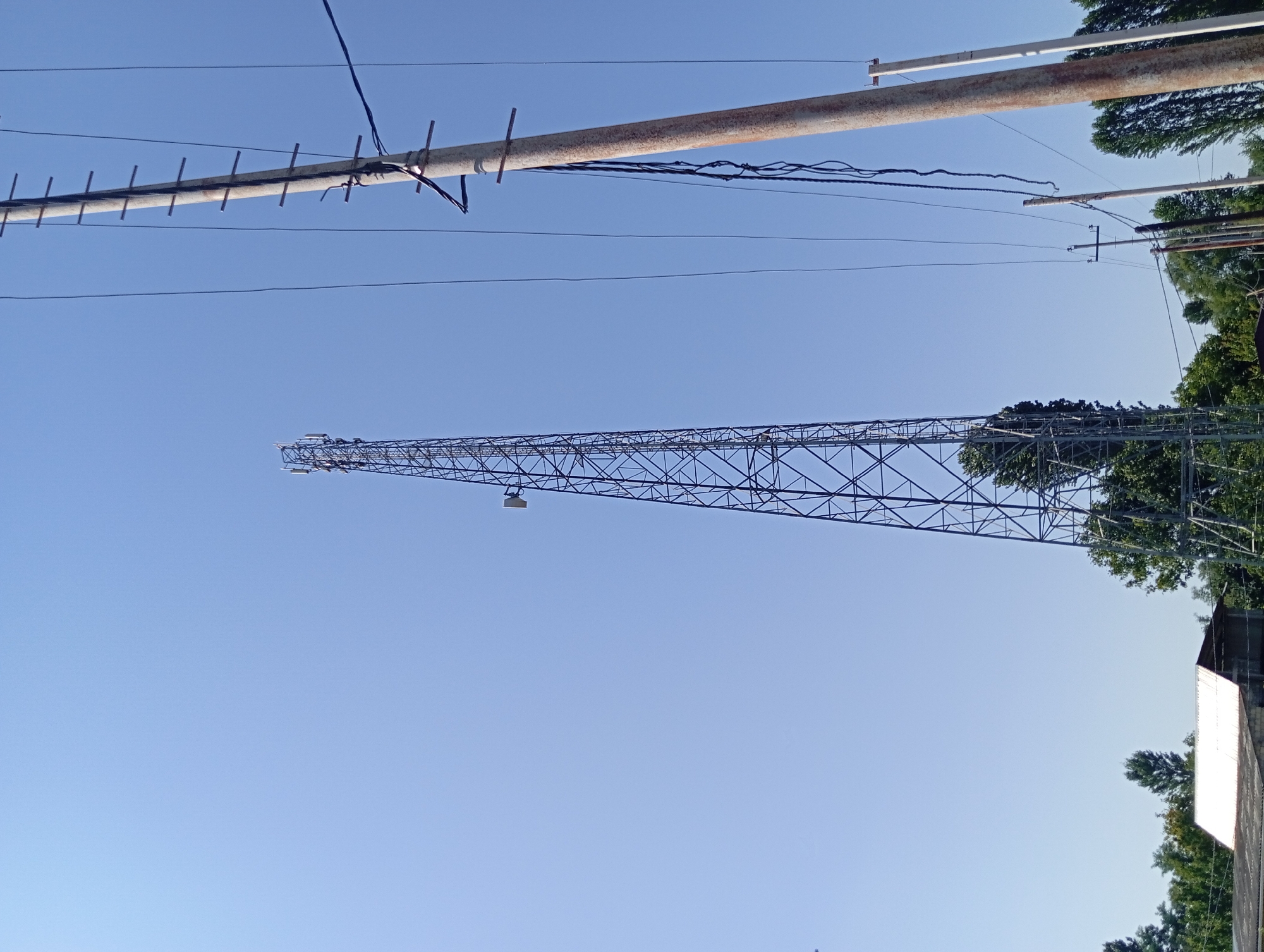A low-angle, full shot depicts a clear blue sky with a prominent cell tower and a utility pole. The cell tower, a tall, lattice structure with antennas, stands to the left of the frame. To the right, a weathered utility pole is adorned with multiple wires and what appear to be ladder rungs. In the lower portion of the image, a white-roofed building with a dark-pitched roof is visible, partially obscured by trees. The environment suggests an outdoor setting, possibly on the outskirts of a settlement, given the presence of the building and scattered trees against the backdrop of the sky. The image is static and captures a typical scene of urban infrastructure.