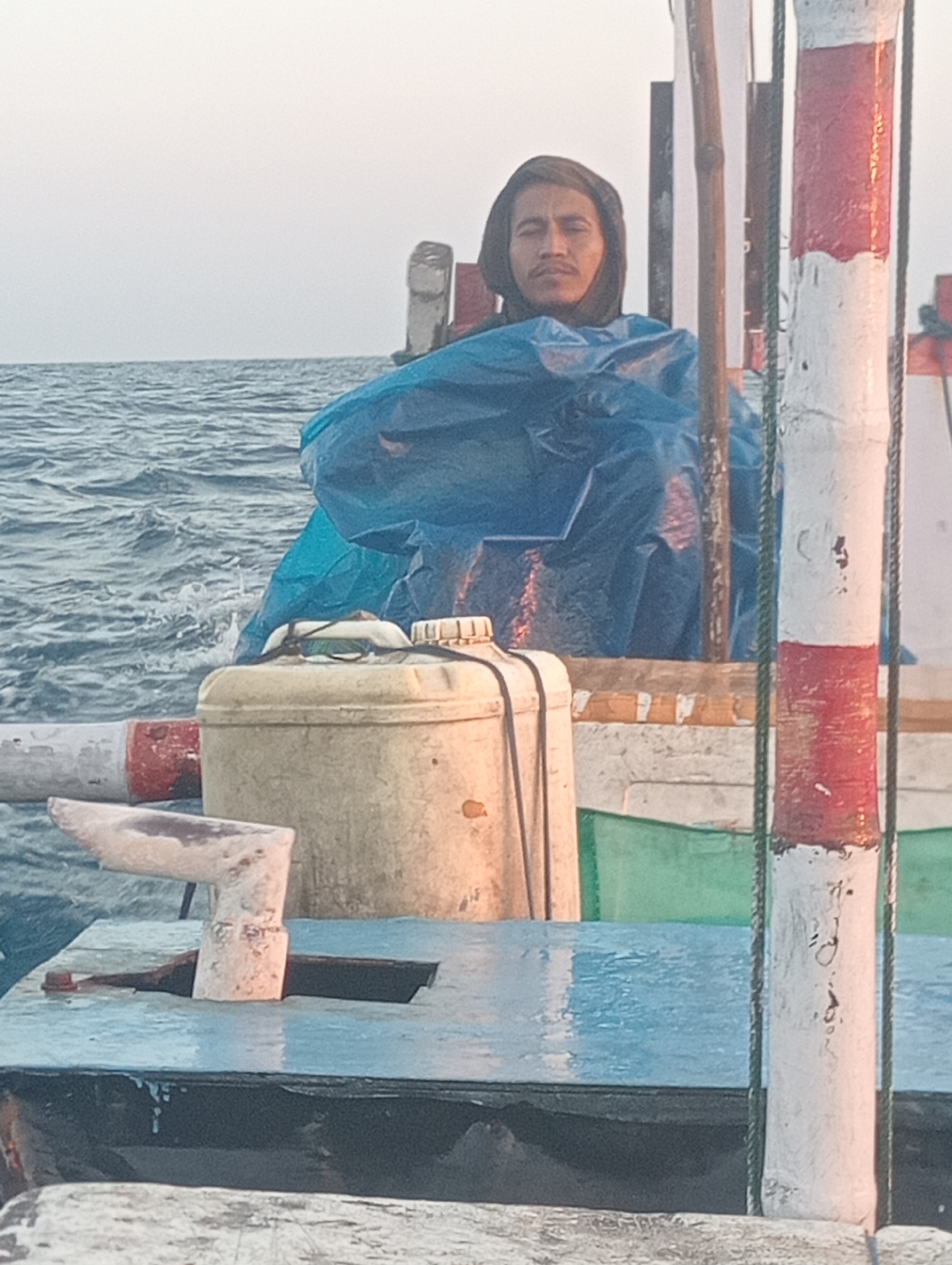 The image shows a person wearing a dark hooded garment sitting on a boat in the ocean. The person is partially covered by a blue tarp. In the foreground, there is a white container with a black lid, and a white cleat attached to a wooden surface. A red and white striped pole is visible on the right side of the image. The water is choppy, indicating a rough sea. The sky is overcast and light in color. The setting appears to be at sea, likely off the coast of Pesisir, Indonesia, given the context of the request.