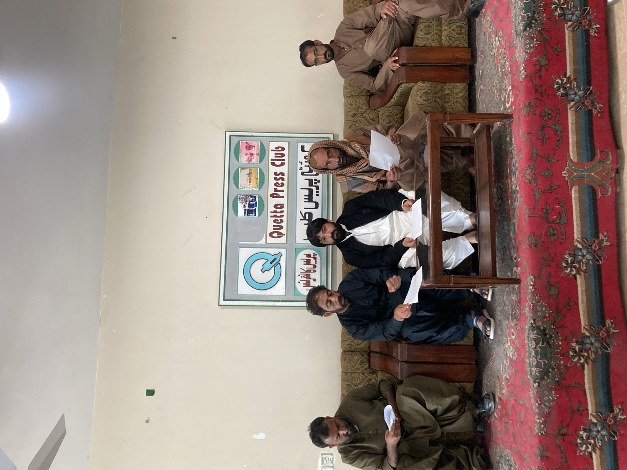 The image depicts a gathering of six men indoors in a location identified as the Quetta Press Club in Quetta City Tehsil, Pakistan, based on the visible sign. The men are seated on sofas and chairs, several holding pieces of paper, suggesting they are engaged in a discussion or meeting. A table is positioned in the center of the group. The background features a wall with the press club's sign and a portion of the ceiling with a ceiling fan. A red rug is on the floor. The men are dressed in traditional clothing.