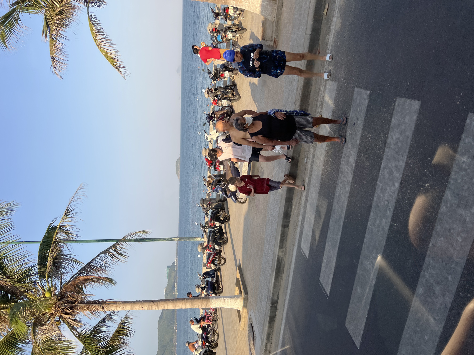 The image depicts a beachfront scene in Nha Trang, Vietnam, with a pedestrian crossing in the foreground. On the promenade behind the crossing, a group of people are gathered near parked motorcycles. A young boy in a red shirt and dark shorts walks across the pedestrian crossing. To his right, a woman in a black tank top and striped shorts, wearing a face mask, stands next to a man in a white t-shirt and dark shorts. Further to the right, a person in a blue swim cap and patterned shorts, wearing a blue long-sleeved shirt with the word "PASSPORT" printed on it, is looking at their phone. Several motorcycles are parked along the promenade. In the background, the sea is visible with small islands or landmasses in the distance, and palm trees line the area. The sky is clear and blue, indicating a sunny day.