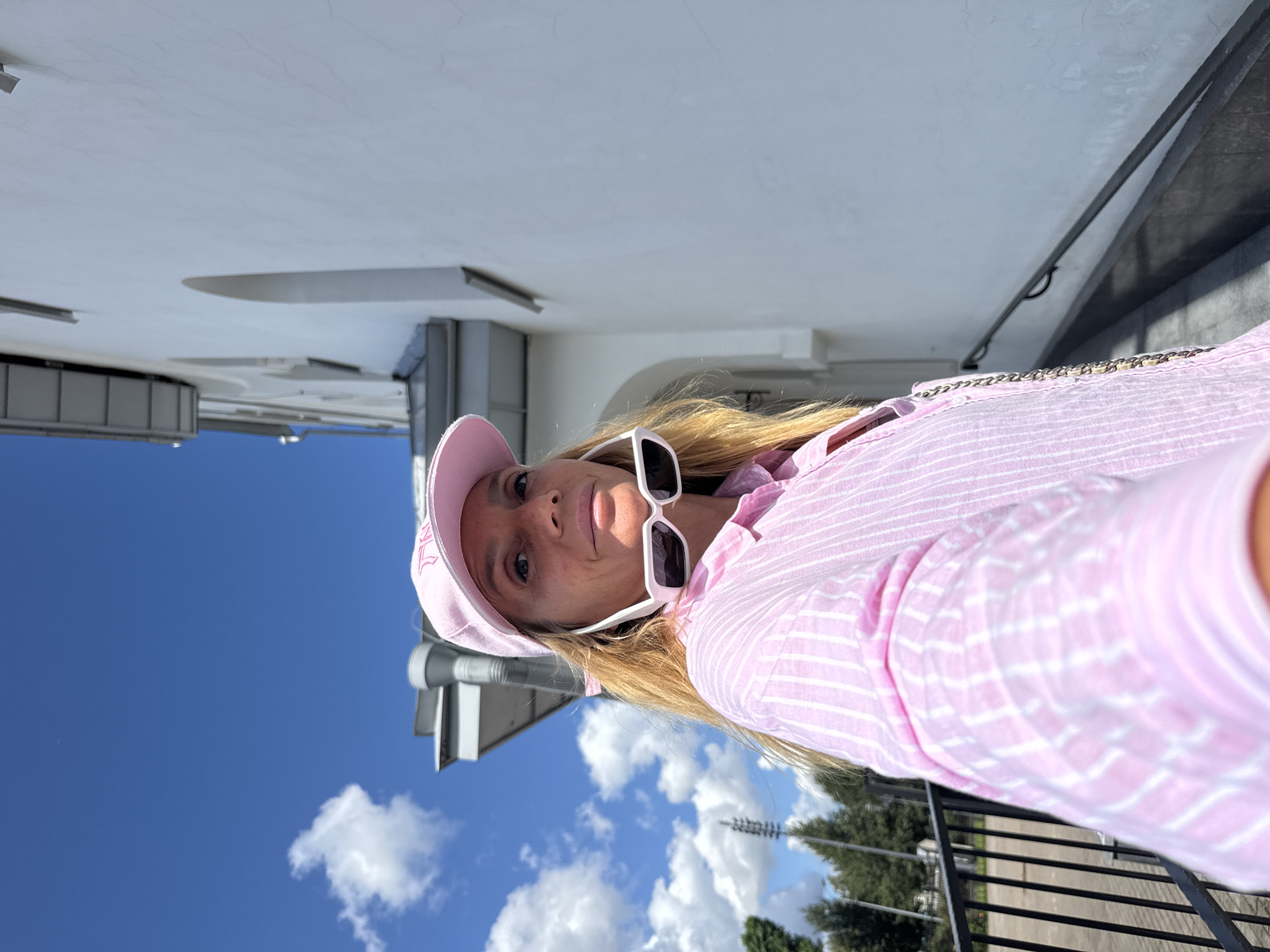 A woman is taking a selfie outdoors, wearing a pink striped shirt, a pink baseball cap, and white sunglasses pushed up on her head. She is standing on a sidewalk next to a white building with arched windows. The sky is blue with scattered clouds, and there are trees and a black metal fence in the background. The location appears to be in a city, with a railing leading up a ramp to the right of the frame.