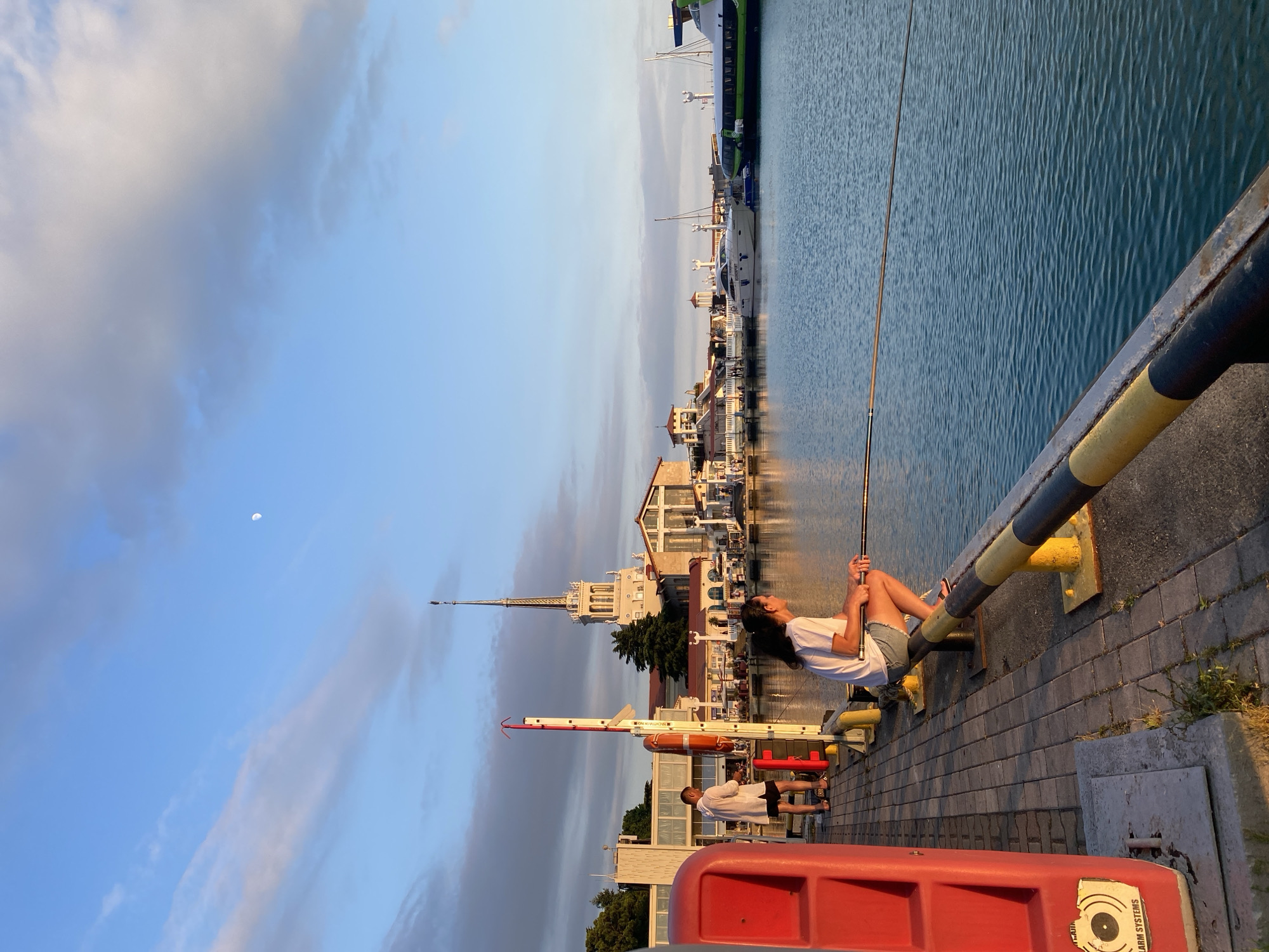 This image depicts a harbor scene in Sochi, Russia, during what appears to be late afternoon or early evening, given the warm, soft lighting and the visible moon in the sky. The sky is a mix of pale blue and soft, wispy clouds, suggesting fair weather.
The foreground is dominated by a paved walkway along the water's edge, with a prominent railing painted in black and yellow stripes. A young woman is seen sitting on this railing, holding a fishing rod and looking up towards the sky with a relaxed expression. She is wearing a white t-shirt and denim shorts. In the mid-ground, another person, a man, is standing and also appears to be fishing. He is wearing a white t-shirt and black shorts. Two large red rectangular objects are visible in the bottom foreground, one of which has text on it: "ALARM SYSTEMS".
The background features a picturesque waterfront with several buildings, including a distinctive tower with a spire. Various boats and yachts are docked in the harbor, with their reflections visible in the calm, rippling water. The architecture suggests a resort or tourist area. The overall atmosphere is peaceful and leisurely, with the individuals enjoying a quiet moment by the water.