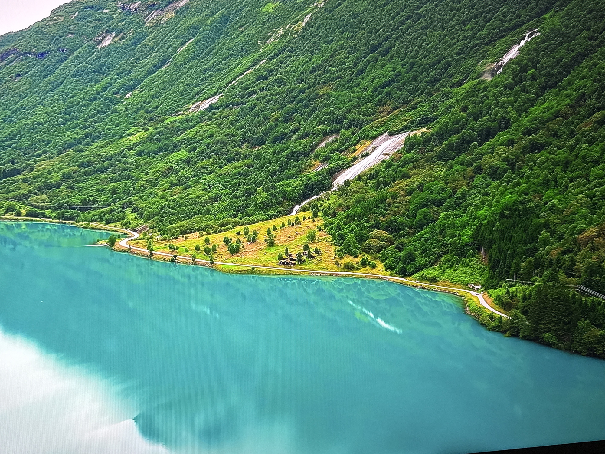 The image shows a vibrant, turquoise lake in front of a lush green, tree-covered mountain. The mountain slopes are steep and dotted with rocky patches and a thin waterfall cascading down from higher up. A winding road runs along the edge of the lake, bordered by a strip of green grass with scattered trees and a few small buildings clustered together. The water reflects the sky, creating a bright, ethereal effect. This scene is not located in the city of Burgas, Bulgaria.