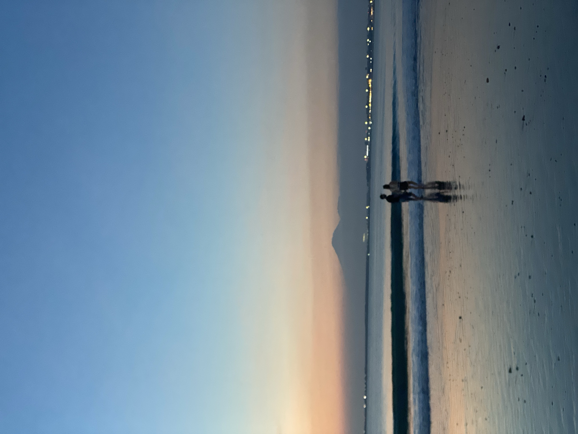 The photograph captures a serene coastal scene at sunset in Kuta Selatan, Indonesia. Two individuals are walking along the shoreline, their figures silhouetted against the wet sand reflecting the ambient light. In the distance, a faintly visible mountain peak rises through the atmospheric haze. A line of lights suggests a distant cityscape or coastal development along the horizon. The sky transitions from a deep blue at the top to a pale orange near the horizon, indicating the setting sun. The overall environment suggests a tranquil evening at the beach.
