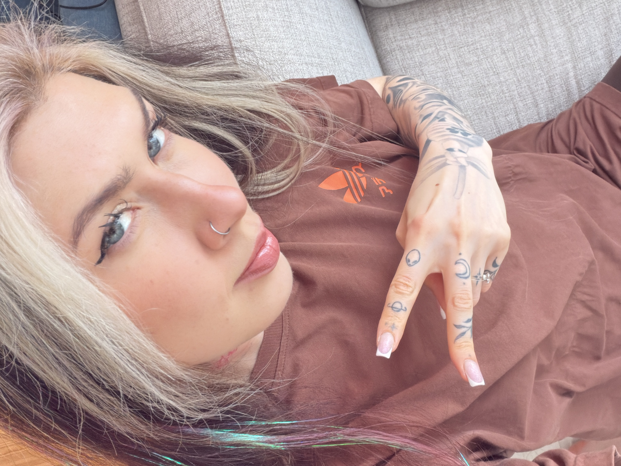 A woman is lying on a light gray sofa, possibly indoors. She is wearing a brown Adidas t-shirt. The woman has fair skin, blue eyes, and light blonde and dark brown hair with streaks of green and pink. She is facing the camera with her head tilted to the left. A silver ring is visible on her left ring finger, and she has a nose ring on her left nostril. Her left hand is raised, displaying finger tattoos, including an alien head, a planet, a moon, and a leaf-like design. Her fingernails are manicured with a french tip. The background consists of a light gray textured sofa. There is no visible information to identify the location as the city of Budva, Montenegro.