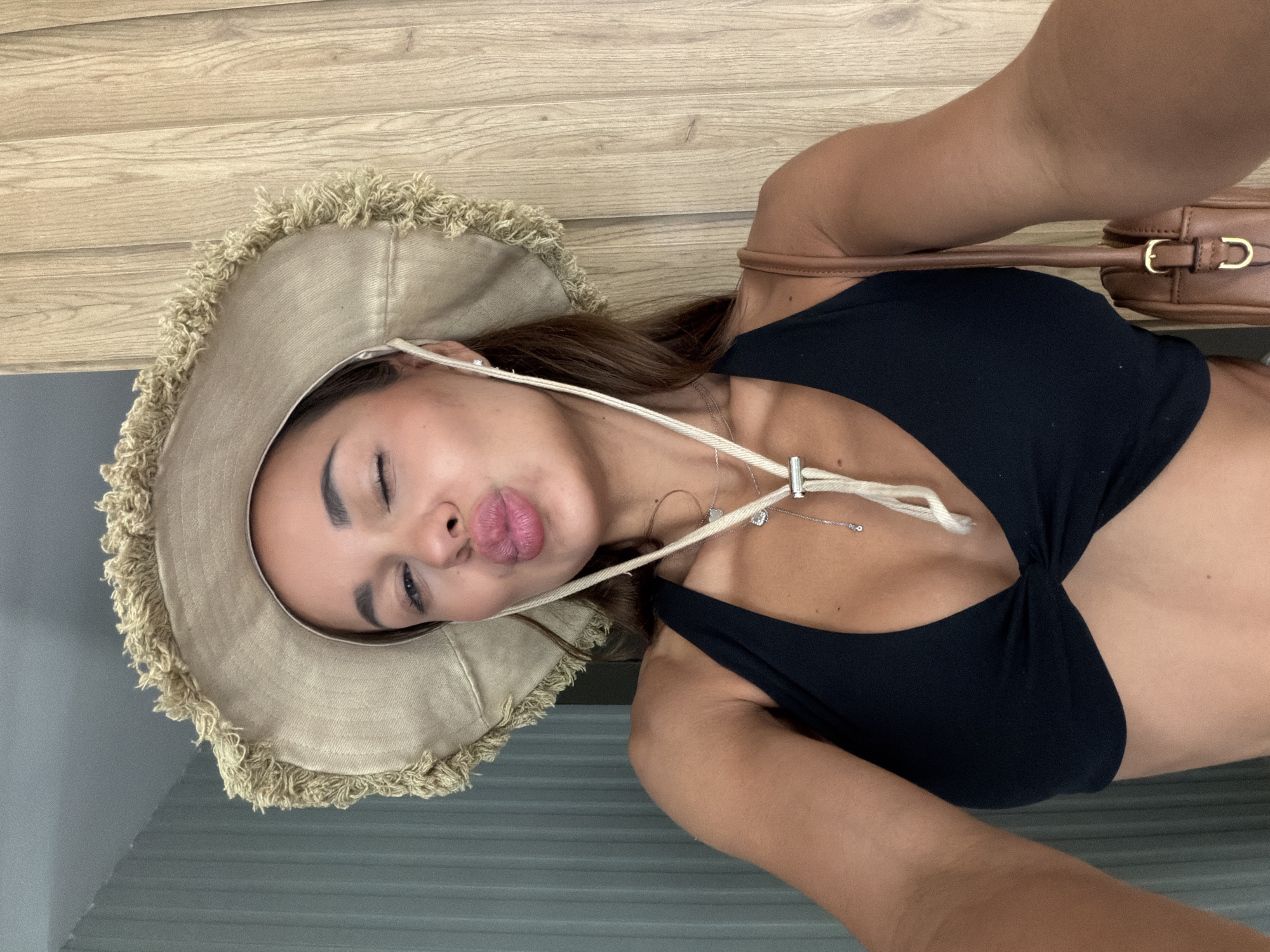 A woman is captured in a close-up, portrait-style shot, wearing a wide-brimmed hat with a frayed edge and a black bikini top. She is looking down, with her eyes closed and her lips puckered in a kiss, as if taking a selfie. A thin necklace with a small pendant is visible around her neck. A brown purse with a gold buckle rests on her shoulder. The background consists of a light-colored wooden surface and a gray striped surface. The image appears to be taken indoors, possibly in a room with wooden paneling. The lighting suggests daytime, but there are no strong indicators of the specific time of day. The overall mood is playful and relaxed. There is no visible text in the image. The location is contextually identified as Bodrum, Turkey.