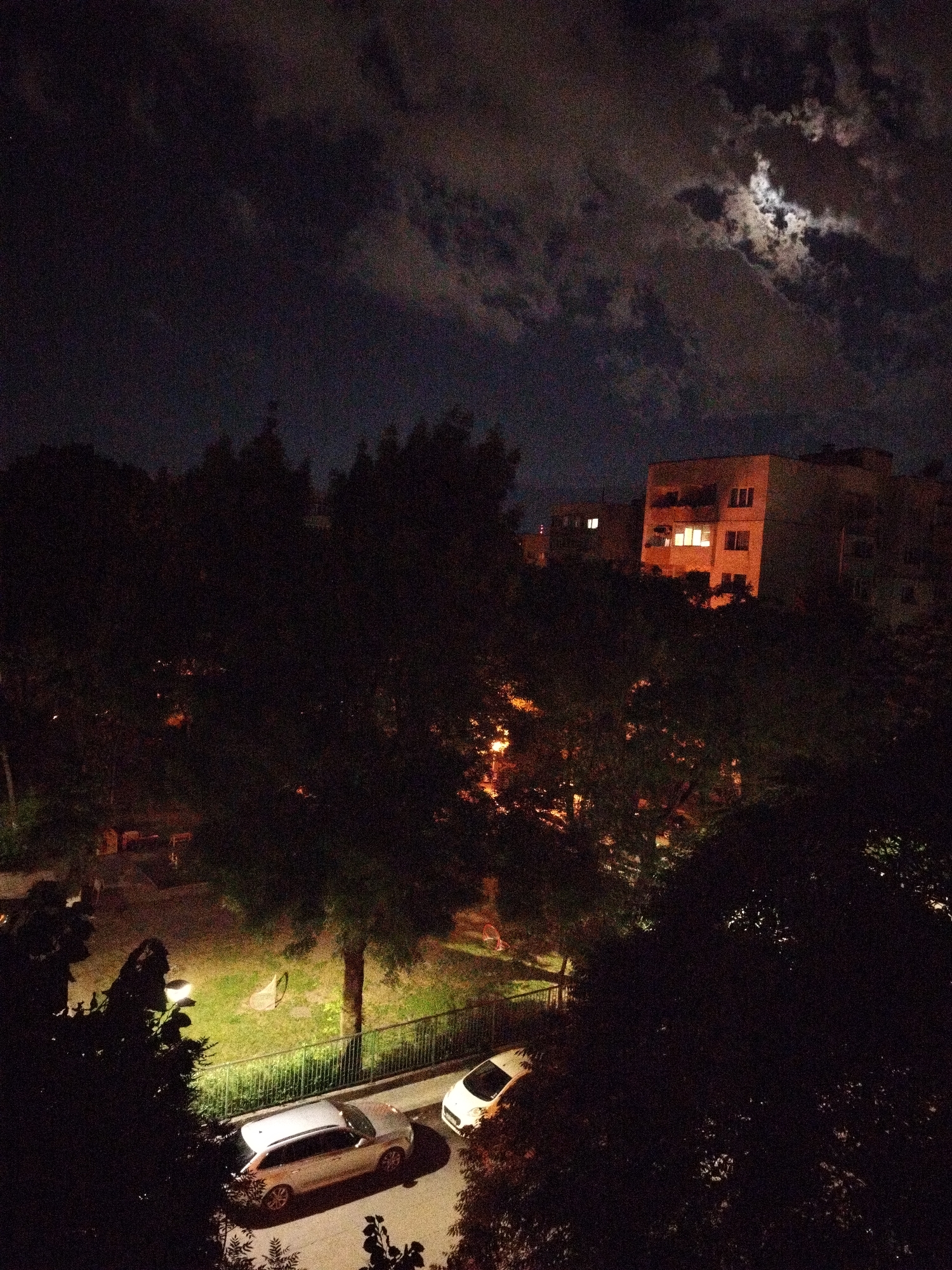 The image depicts a nighttime scene in Sofia, Bulgaria, likely taken from an elevated perspective, such as from an apartment building. In the foreground, two light-colored cars are parked adjacent to a metal fence, illuminated by a streetlight. Beyond the fence, there's a green space, possibly a park or courtyard, with visible trees, grass, and park benches. In the background, a multi-story building with illuminated windows is visible. The sky is dark with scattered clouds and a visible moon. The scene is primarily lit by streetlights, building lights, and moonlight. No people are visible in the frame. The general environment suggests a residential area in Sofia at night.