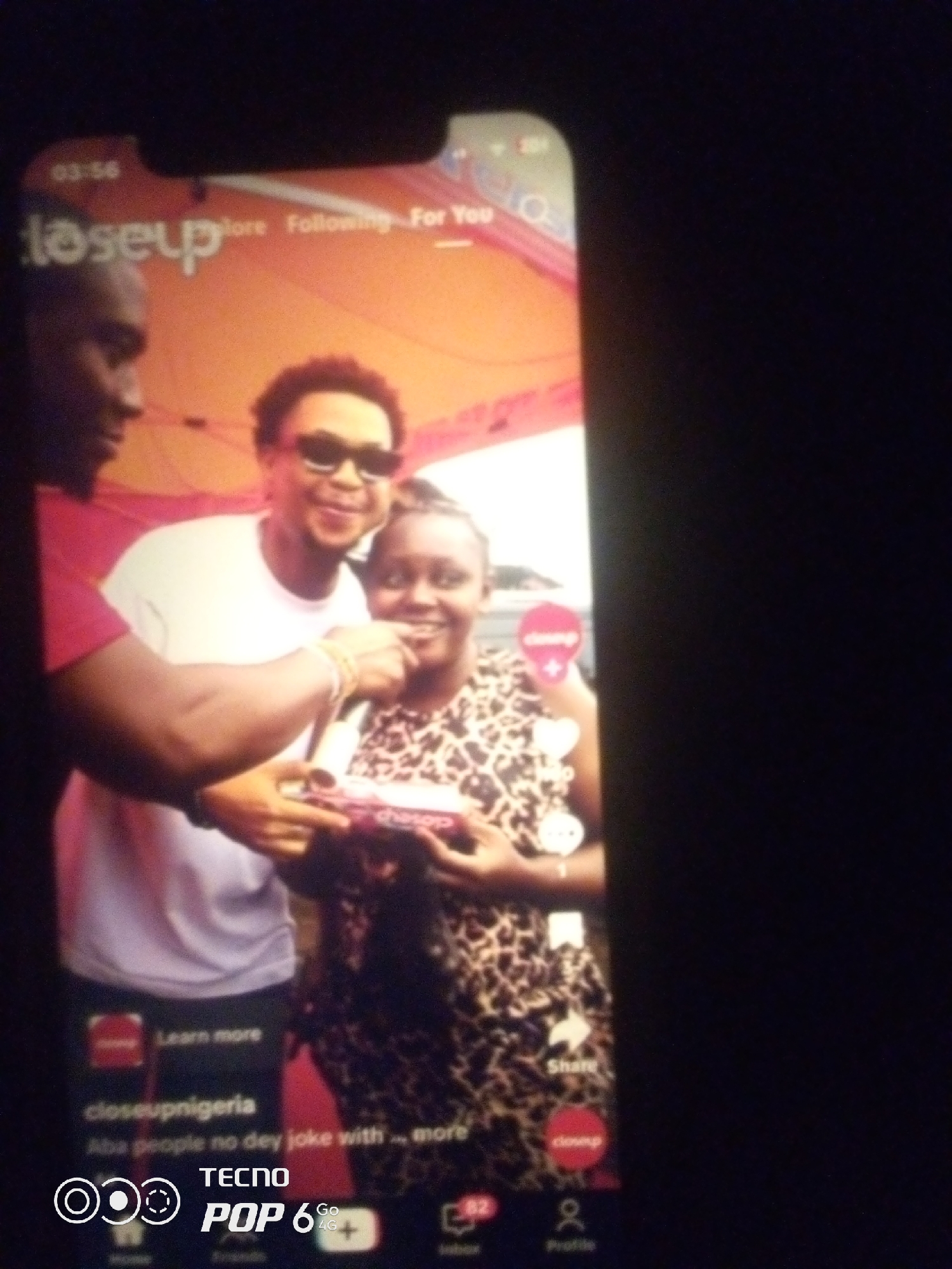 A medium-shot vertical, blurry image shows a screen display of a phone showing a TikTok video. The video displays two individuals: a person on the left with dark skin, wearing a bright pink shirt, and a person in the center with dark skin, wearing sunglasses and a white t-shirt. The person on the right, a woman with dark skin, wears a leopard-print dress and is holding a small pink box with white lettering. The background of the video appears to be an outdoor event with an orange tent and possibly a crowd. Below the video, the phone screen shows interface elements including "Learn more," "closeupnigeria," and caption text that reads "Aba people no dey joke with ... more." At the bottom of the phone screen, brand logos and icons for "TECNO POP 6 GO 4G" are visible. The phone is displayed in a dark environment. The location of Howayi, Nigeria, is not depicted in this image.