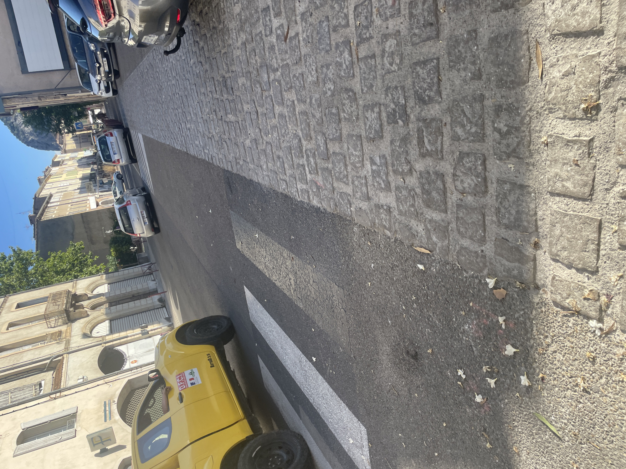 A yellow utility truck with "Barum 2.5" on the side is parked on a street, partially blocking a pedestrian crossing. The truck has stickers on its door including a French flag and a logo with the text "D.F.C.I.". Several cars are parked or driving on the street. The street is lined with buildings and a cobblestone sidewalk. The sky is clear and blue.