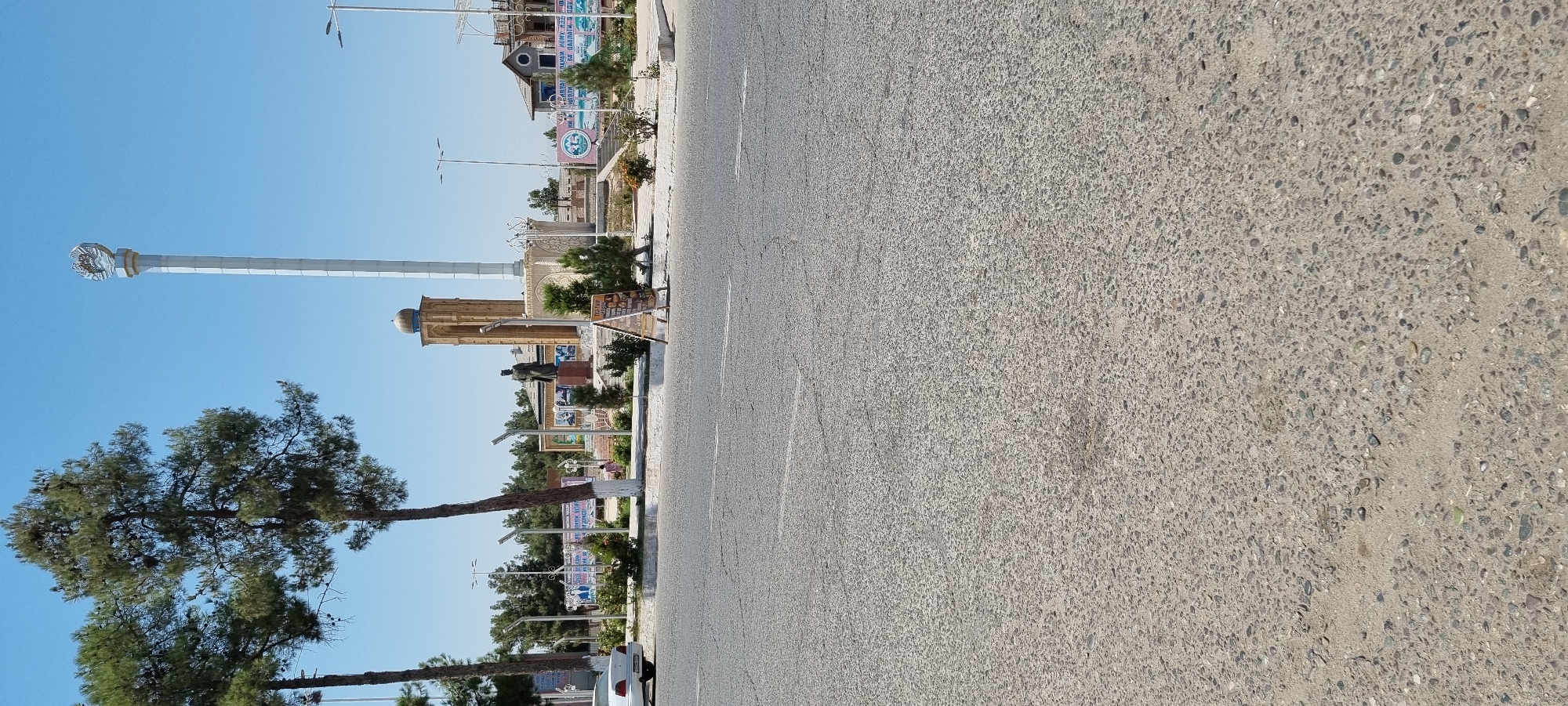 The image shows a wide asphalt road in the foreground with visible cracks and some loose gravel. In the background, a public square is depicted with several key features. A tall, white monument topped with a circular emblem stands prominently. To its left, a minaret-like structure with a dome can be seen. In front of these, a statue of a person in a formal pose is visible. The square is bordered by trees, bushes, and some decorative fencing. Several banners with text are displayed, and a few people are scattered throughout the area, including a person near a white car on the left side of the road. An A-frame sign displaying food items is placed on the sidewalk. The sky is clear and blue. The overall scene suggests a public space within a city, likely a monument or memorial area in Ismoili Somoni, Tajikistan.