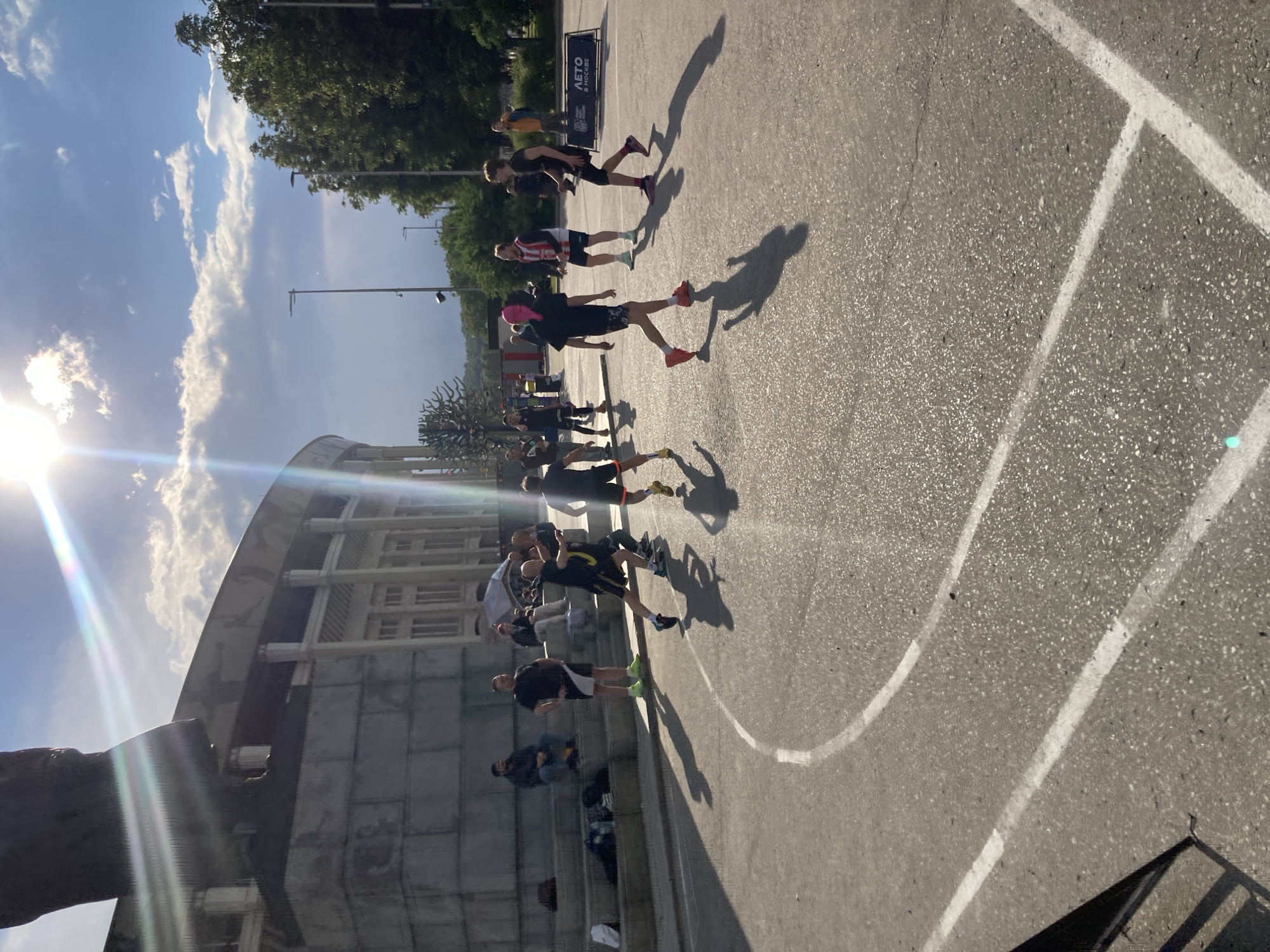 The image depicts a group of people on an asphalt surface with white lines, likely a sports area or park in Moscow, Russia. The presence of a sign that reads "ЛЕТО в МОСКВЕ" confirms the location as Moscow. The individuals are mostly young and appear to be engaged in a casual stroll or light exercise. They are wearing casual clothing and sneakers, and their shadows are cast long due to the low sun angle, indicating either early morning or late afternoon. To the left, there's a portion of a building, possibly a stadium or sports complex, and some people sitting on steps. The weather appears sunny with some clouds in the sky.