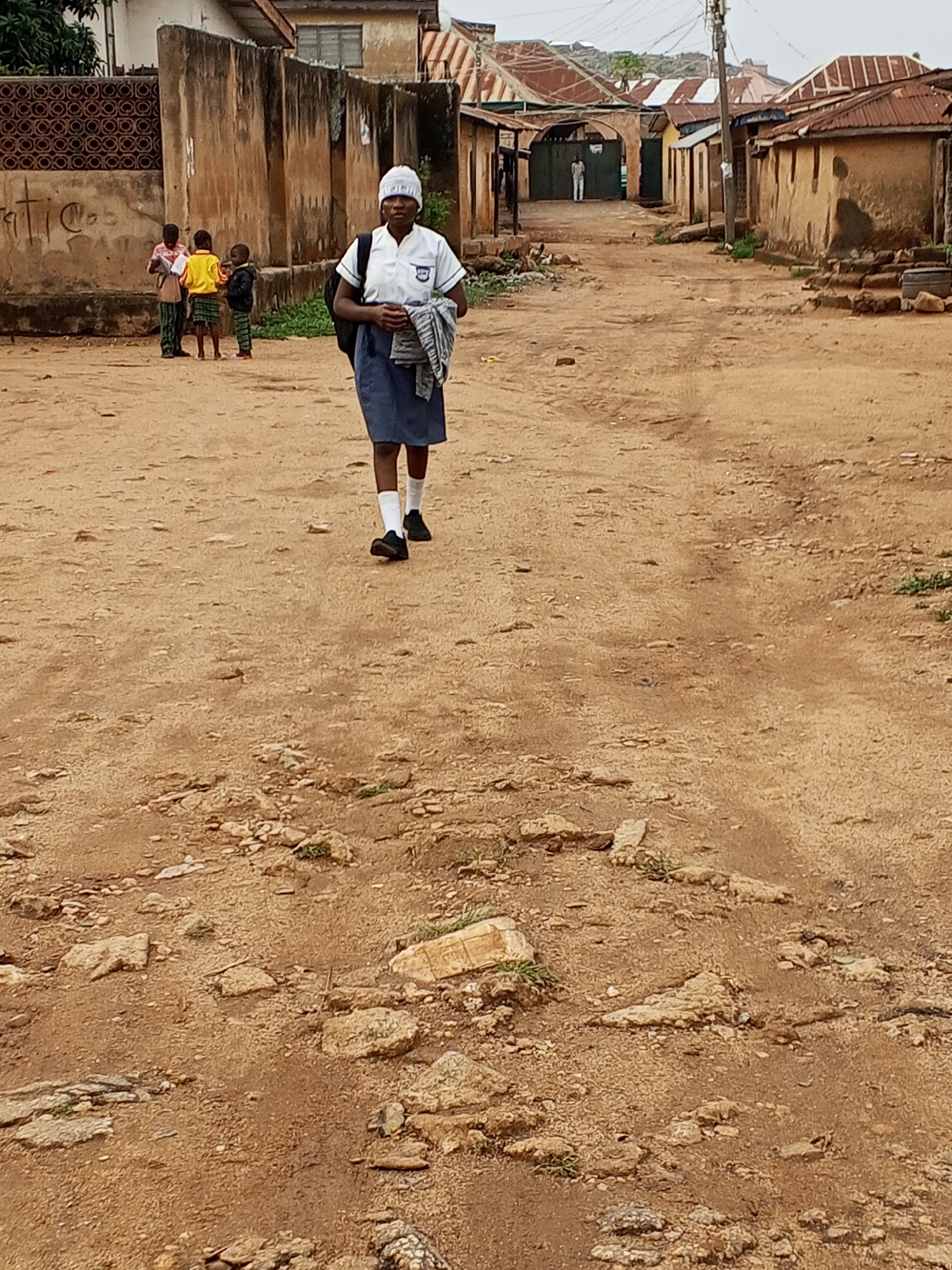 The image shows a young woman walking down a dirt road in Jos, Nigeria. She is wearing a school uniform: a white short-sleeved shirt, a navy blue skirt, white socks, and black shoes. She also has a white head covering and a black backpack. In her right hand, she is holding a rolled-up garment, likely a sweater.
In the background, to the left, are three young children. The child closest to the viewer is wearing a yellow shirt, a green plaid skirt, and sandals.
The road is uneven, with exposed rocks and patches of bare soil. On either side of the road are dilapidated buildings with corrugated metal roofs. In the distance, a figure dressed in white stands in an archway, and electric poles and wires are visible against a hazy sky.