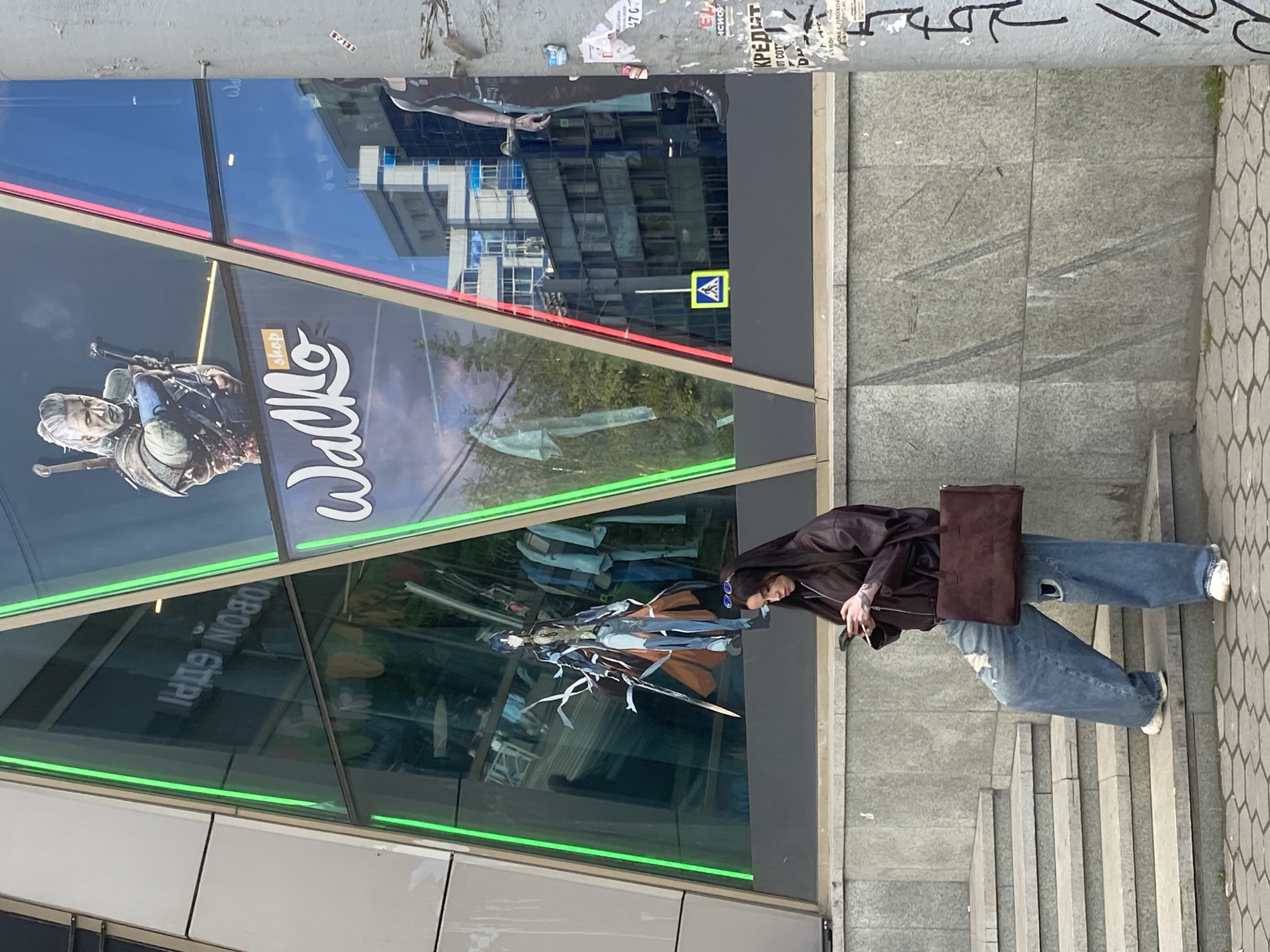 The photo captures a scene in Yekaterinburg, Russia, in front of a building featuring a shop sign displaying "Wallko." The sign showcases promotional material related to the video game "The Witcher" and "Genshin Impact," and reflects a cityscape within its glass. A person is standing on a set of stairs next to the building, with long dark hair, wearing a dark jacket, ripped blue jeans, white sneakers, and carrying a brown handbag. The person is holding a cigarette in their hand. The wall adjacent to the building has various posters and graffiti. The sidewalk is composed of hexagonal-shaped paving stones.