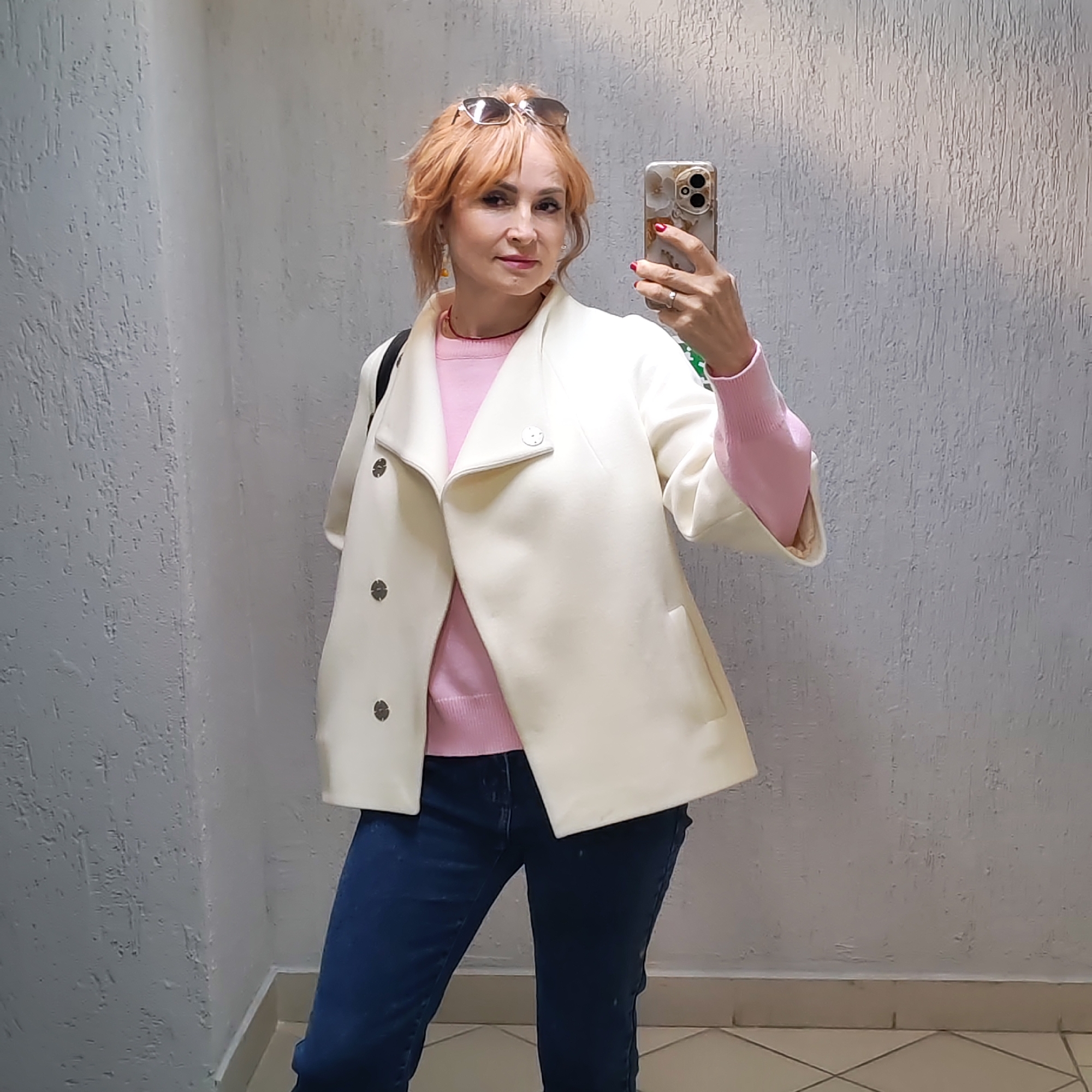 A woman is taking a selfie indoors, seemingly within a building in the city of Chaikovsky, Russia. The woman has short, reddish-blonde hair and is wearing sunglasses on her head. She is dressed in a pink sweater, dark blue jeans, and a cream-colored jacket with large buttons. She is holding a mobile phone in her right hand, pointed towards herself to capture the image. The background consists of textured, gray walls and a tiled floor. The lighting in the space appears soft and diffused.