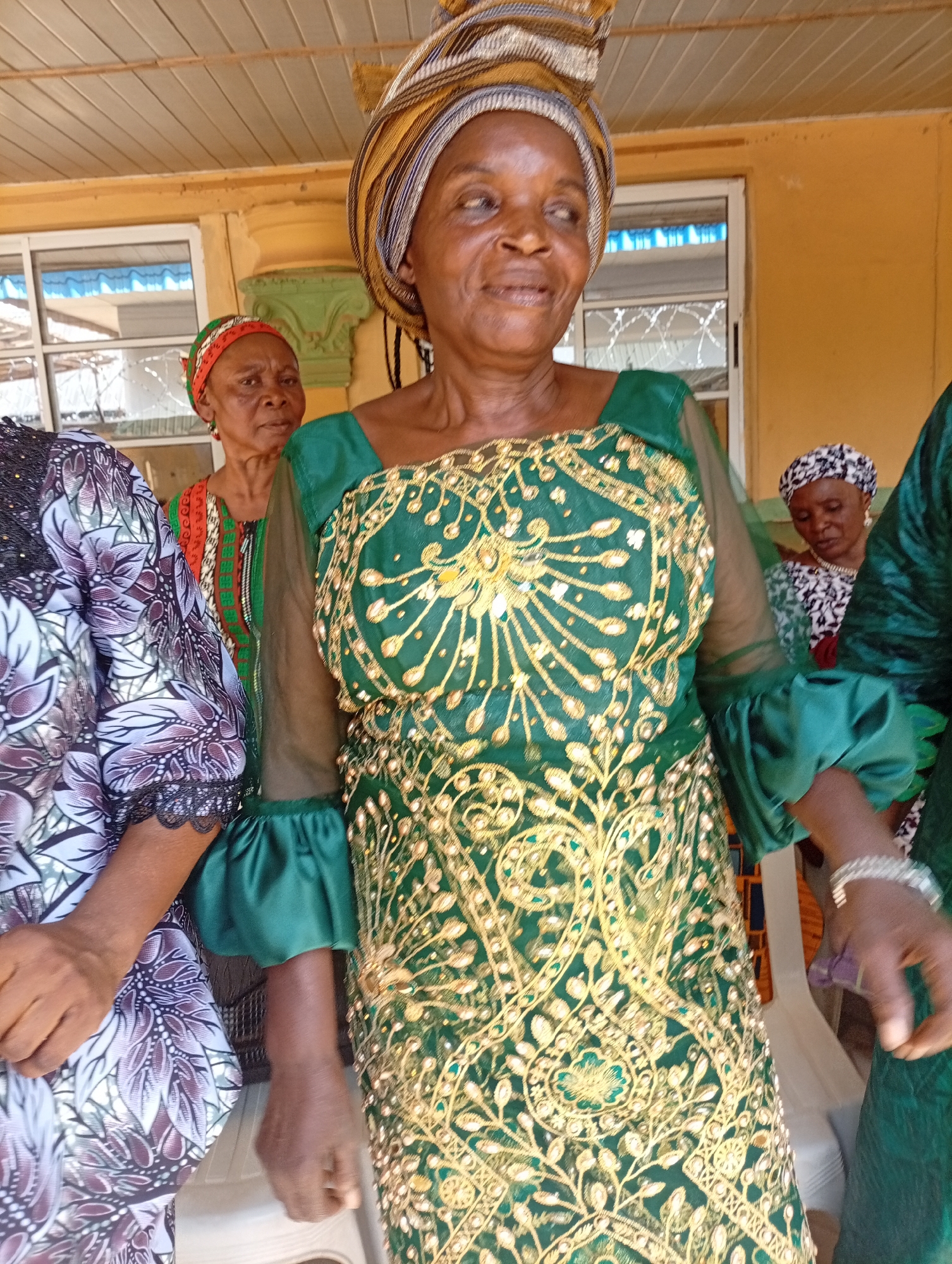 The image shows a group of women, likely in Rido, Nigeria, dressed in traditional attire. The woman in the foreground wears a green dress with gold embroidery and a patterned head wrap. Behind her, other women are visible in similarly vibrant clothing. The setting appears to be outdoors, possibly near a building with yellow walls and window frames.