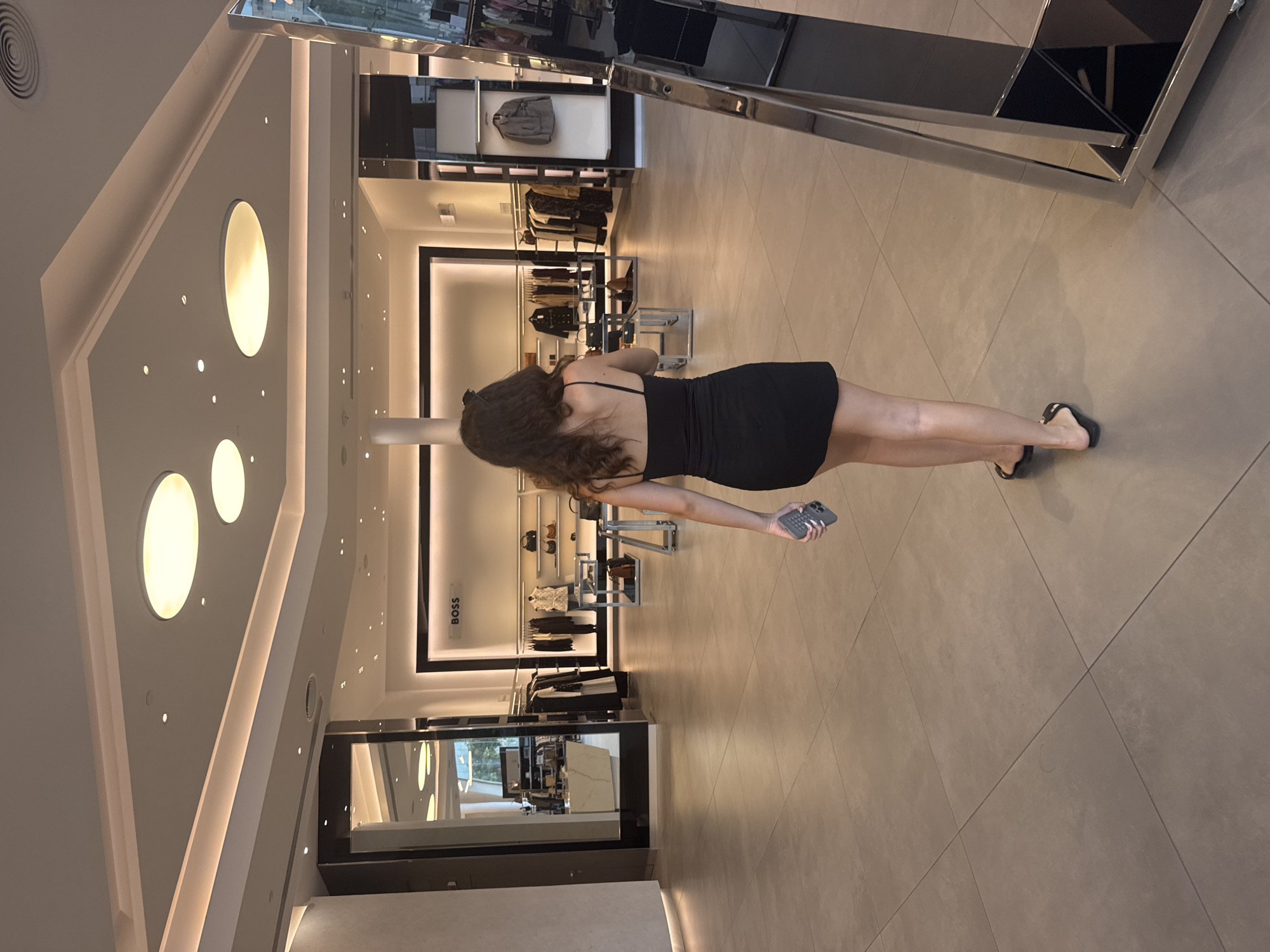 A woman is walking away from the camera in a clothing store. She is wearing a black spaghetti strap dress and black flip-flops. She is holding a gray mobile phone in her right hand. The store appears to be a high-end boutique, with clothing racks and display shelves visible in the background. The floor is tiled in a light gray color. The lighting is bright and modern, with recessed ceiling lights and accent lighting. The store has a minimalist design, with clean lines and neutral colors. The woman is the sole subject in the foreground. The background shows racks of clothing, handbags, and a prominent "BOSS" logo on the wall. The overall impression is one of a modern, well-organized retail environment. The store is located in Limassol, Cyprus, as indicated by the presence of the BOSS store.