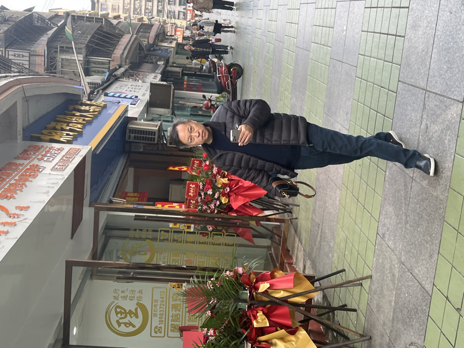 The scene is set on a tiled sidewalk in a commercial area of Jiangjin, China, under overcast lighting conditions. A middle-aged East Asian male occupies the central foreground, walking towards the viewer from the right side of the frame. He wears a dark puffer jacket, a dark long-sleeved shirt, dark blue jeans, and black slip-on shoes. In his right hand, he holds a silver-colored smartphone, with his gaze directed at its screen. His left hand holds a dark shoulder bag with a brown strap. His facial expression is neutral.
To the immediate left of the central figure, a storefront with large glass windows and doors displays gold Chinese characters, including "理" and a telephone number, "18983726413". Outside this establishment, several large floral arrangements, decorated with red and gold ribbons and featuring green, palm-like foliage, are positioned on metal stands, suggesting a recent opening or celebratory event. Further down the street to the right, other pedestrians are visible, one next to a parked red electric scooter. The street is lined with multi-story buildings, displaying various shop signs and external air conditioning units. The sidewalk is paved with a pattern of alternating grey and light green rectangular tiles.